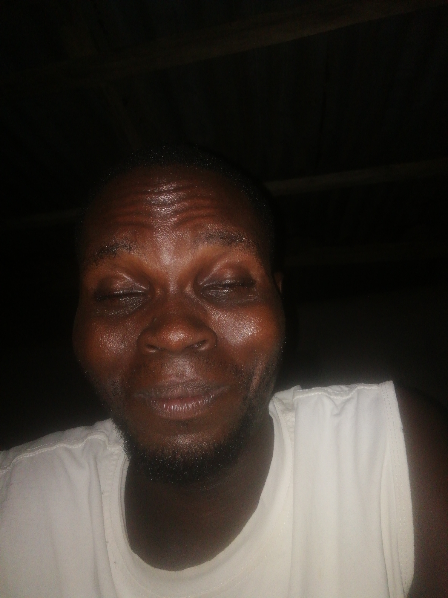 A close-up photograph depicts a man with dark skin, wearing a white tank top. His eyes are closed. A short beard is visible on his face. The background is dark. There is no contextual information in the image to definitively determine that the location is Ikorodu, Nigeria.
