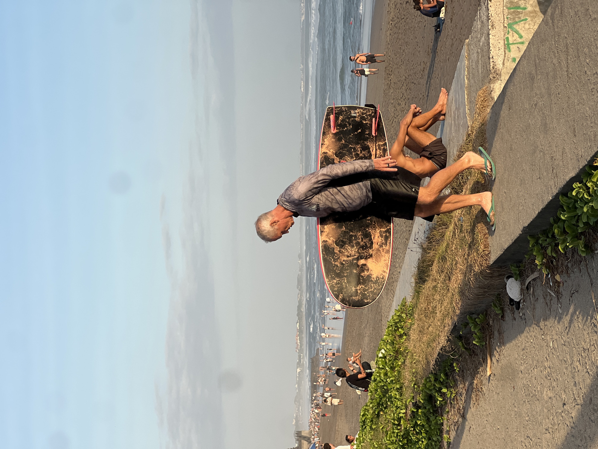 An eye-level, medium shot captures a dynamic scene on the coast of North Kuta, Indonesia. The foreground is dominated by a concrete walkway bordering lush green vegetation. On the walkway, a fair-skinned man with grey hair, wearing a grey patterned long-sleeved rash guard and black shorts, walks from right to left, carrying a surfboard with an orange and black marbled design. His left foot is adorned with a green flip-flop. Seated on the concrete wall to his right, facing away from the camera and partially obscured by the surfer, is another fair-skinned person in black shorts, their legs crossed.
Beyond the walkway and wall, a sandy beach stretches out, populated by numerous individuals. Some are standing, others are sitting, and a few are walking along the shoreline. The ocean beyond is active with moderate waves, suggesting a surfing spot. The sky above is a pale blue with scattered, wispy clouds. In the distance, a town or resort area is visible along the coastline, characterized by buildings and a hazy skyline. The overall impression is one of a lively beach community enjoying the late afternoon or early evening.