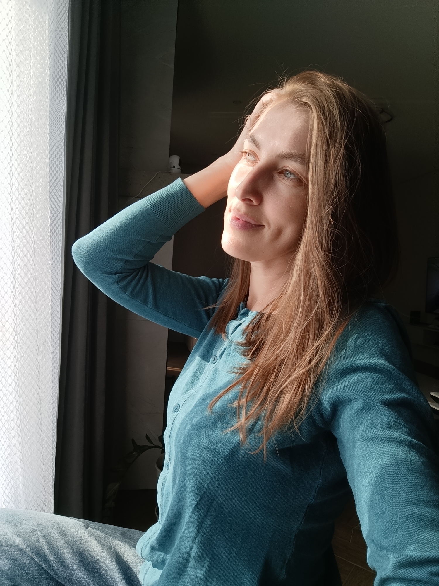 A woman with light brown hair and blue eyes is looking out of a window. She is wearing a teal-colored button-up shirt and matching pants. Her left hand is resting on her head. The window has white curtains and a dark gray curtain behind them. The location is in a room with natural lighting and a view of the outside. It's unknown if this image was taken in Chaikovsky, Russia.