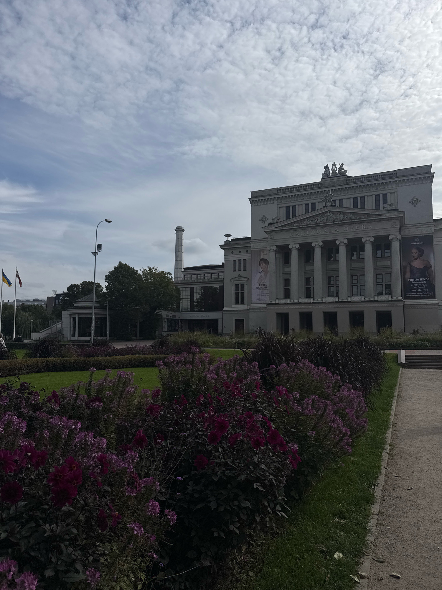 The image shows the National Opera in Riga, Latvia, on a cloudy day. In the foreground, a well-maintained flower bed filled with vibrant purple and pink flowers runs along a pathway. Behind the flowers, a green lawn stretches towards the building. The National Opera, a grand neoclassical structure, dominates the background. It features a large portico with towering columns, and statues are visible on the roof. Banners advertising upcoming events are displayed on the facade. To the left of the opera house, a distinctive cylindrical chimney rises from a lower building. In the distance, three flags fly on poles: the Ukrainian flag, the Latvian flag, and an unidentifiable third flag. A streetlamp stands near the flags, and trees are scattered throughout the scene. The overall impression is one of an important cultural institution set amidst a carefully landscaped urban park.