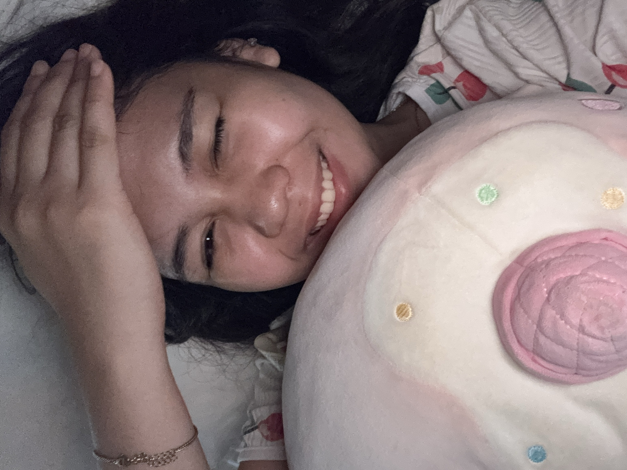 In Davao City, Philippines, a female individual is captured in a close-up, horizontal portrait, lying down and smiling broadly with her right eye closed in a wink. Her left arm is extended overhead, resting on a light-colored surface. She has dark hair, and her complexion appears moist or shiny, possibly from low light conditions. She is wearing a white shirt featuring red and green fruit-like patterns (possibly cherries). The individual is embracing a large, pale pink and white plush toy, resembling a donut, with a prominent pink quilted center and various small, colorful embroidered circular patterns (yellow, blue, green) across its surface. The background consists of dimly lit, indistinct light-colored fabric or surfaces.