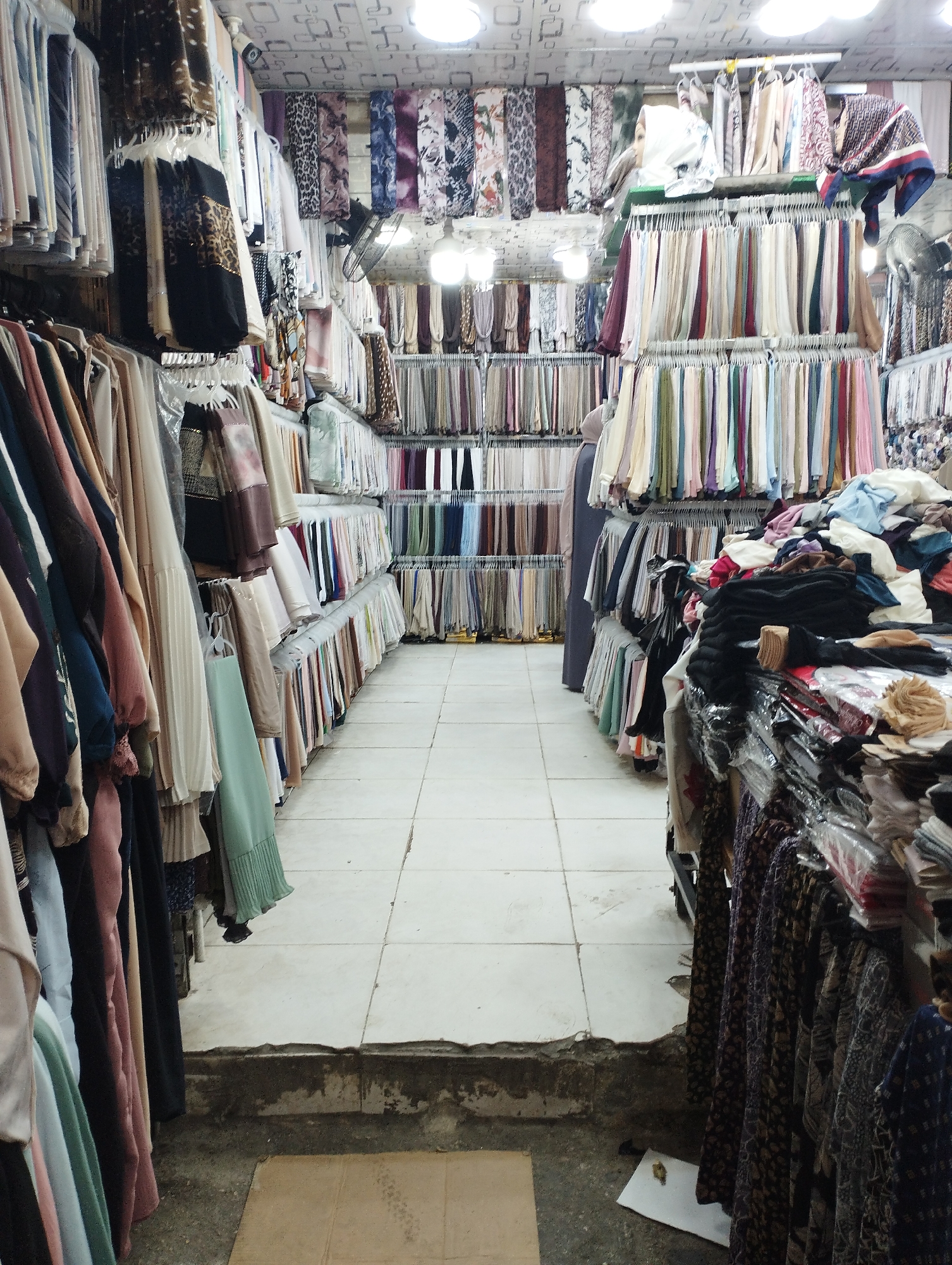 The image depicts an indoor market stall in Baqa'a camp, Jordan, filled with a variety of textiles. Racks of colorful scarves and fabrics are displayed prominently, organized by color and pattern. Some are draped, while others are neatly folded on shelves. A few mannequins, partially visible, are also dressed in the displayed garments. The stall appears to be a retail space with tiled flooring and overhead lighting. The overall impression is one of abundance and variety in textile merchandise.