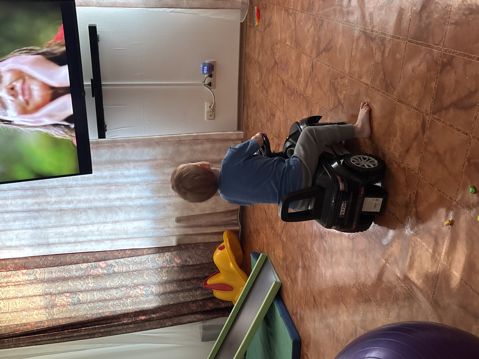A young child with blonde hair is sitting on a black toy car on a tiled floor indoors. The child is wearing a blue shirt and gray pants. A television showing a woman's face is on the left side of the image. In the background, there are patterned curtains, a child's slide, a yellow toy, a purple exercise ball, and an electronic device plugged into the wall. The location is ферма, Russia.