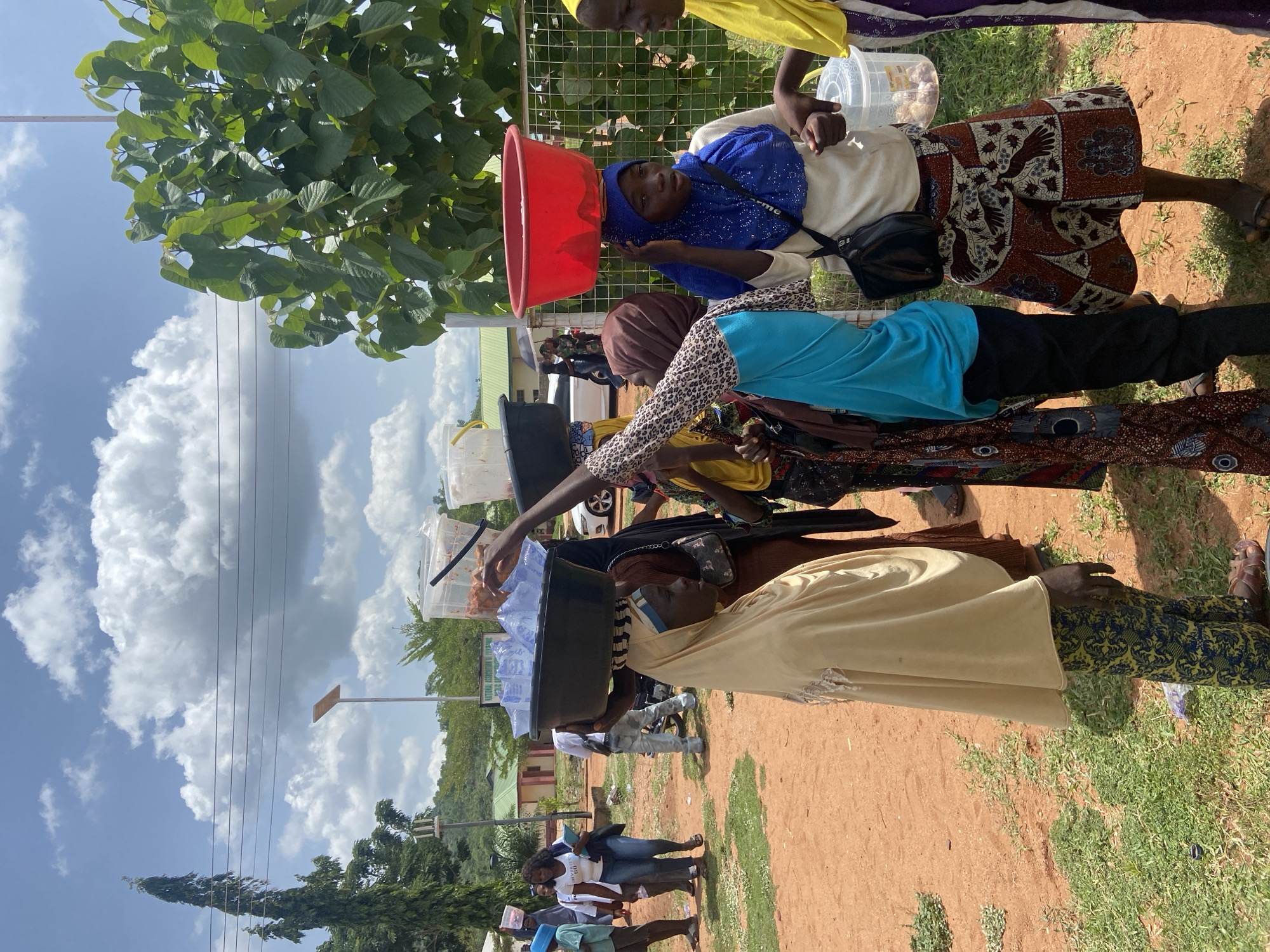 In Lokoja, Nigeria, a group of people are seen on a dirt ground near a grassy area and a building. Several individuals are carrying items on their heads, including containers with food and water. One person is wearing a red bucket on their head. There are other people standing and walking in the background. The sky is visible with white clouds.