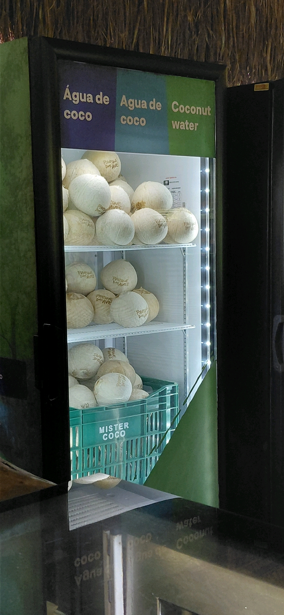 The image shows an open refrigerator displaying a large quantity of whole coconuts. The refrigerator has a green exterior and a black door frame. Above the coconuts, there is a sign with text in three colored sections: purple, blue, and green. The purple section reads "Água de coco", the blue section reads "Agua de coco", and the green section reads "Coconut water". The coconuts are arranged on shelves within the refrigerator. Some of the coconuts have branding written on them, which appears to be "PARQUE DAS AVES". In the lower section of the refrigerator, a green plastic crate labeled "MISTER COCO" is visible, also filled with coconuts. The background is dark, and suggests an indoor setting, possibly a market or a store. There are no people visible in the image. The scene appears to be from a retail establishment in Foz do Iguaçu, Brazil, given the presence of "PARQUE DAS AVES", which is a well-known tourist attraction in that city.