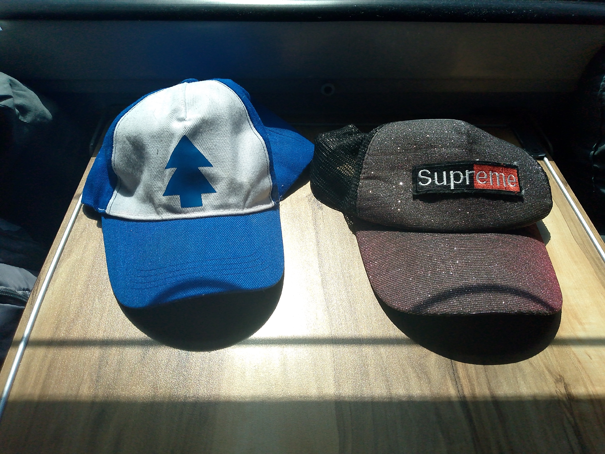 Two hats are placed on a wooden surface. The hat on the left is blue and white with a blue pine tree symbol. The hat on the right is black with a glittery texture and a "Supreme" logo patch. The hats are positioned side-by-side, casting shadows on the surface. The background is dark and out of focus.