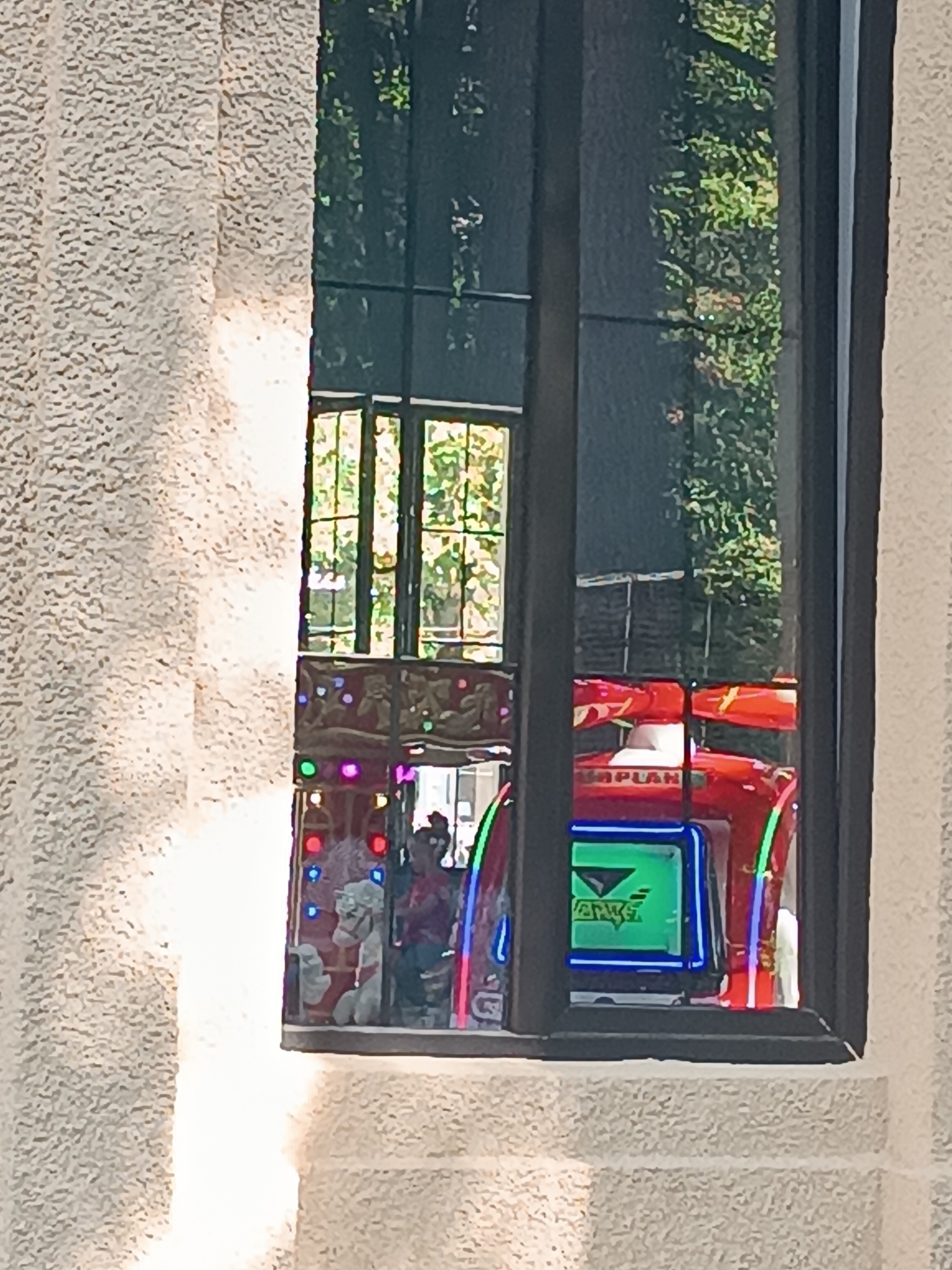 The image shows a reflection in a window of what appears to be an arcade or amusement area. In the reflection, a red amusement ride, possibly a car-themed ride, is visible with a green screen displaying a logo. Lights, possibly from the ride or the surrounding area, are also visible. Part of a carousel with figures and lights is also reflected. The exterior of the window frame is made of textured, light-colored stone. This scene is located in the city of Borjomi, Georgia.