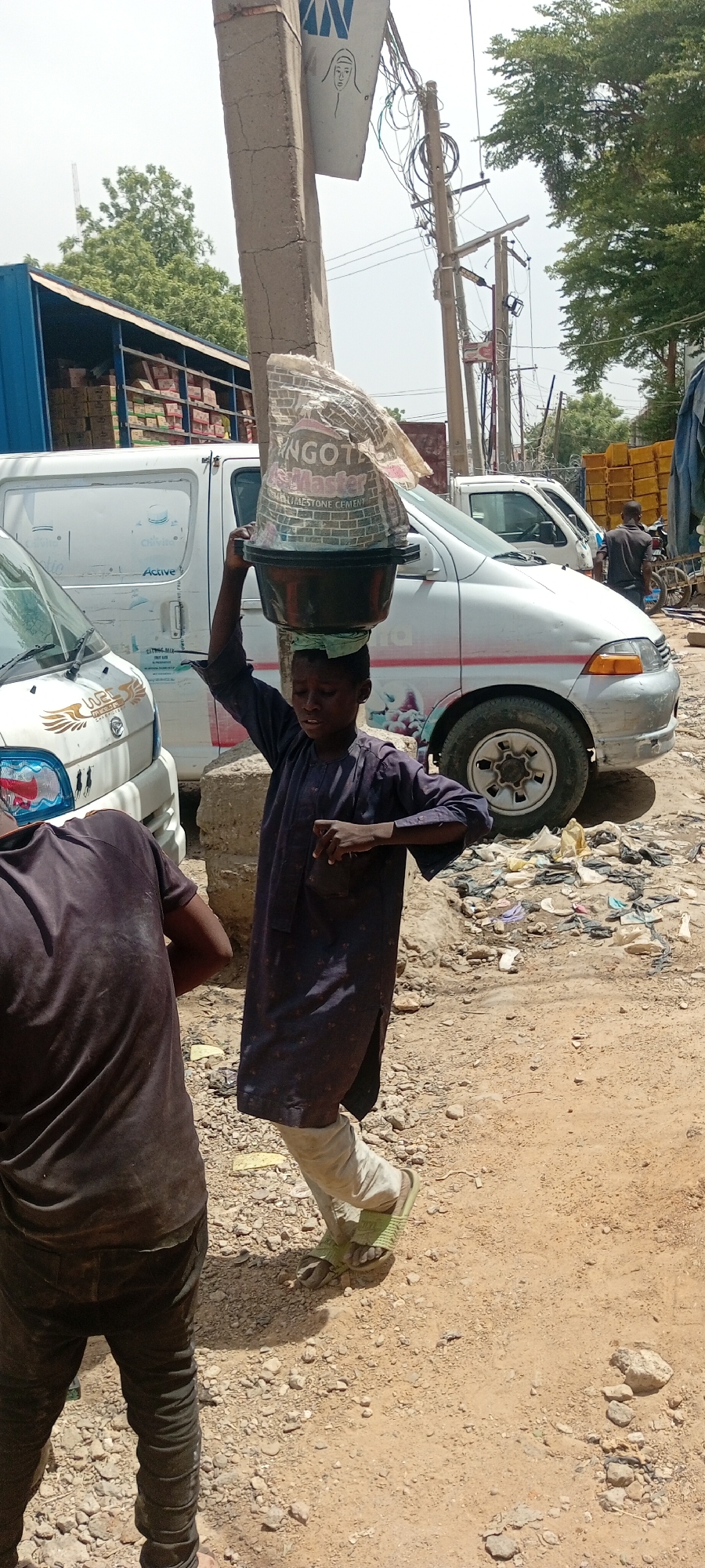 In Fagge A, Nigeria, a young person carries a large bag of Dangote limestone cement atop a black basin on their head. The person is wearing a long-sleeved, patterned purple robe, white pants, and green sandals. The person is walking on a dirt and gravel surface littered with trash. Behind the person, a white van is visible, along with other parked vehicles. A man in a dark shirt stands to the left of the person carrying the cement. Overhead power lines and a utility pole are also visible.