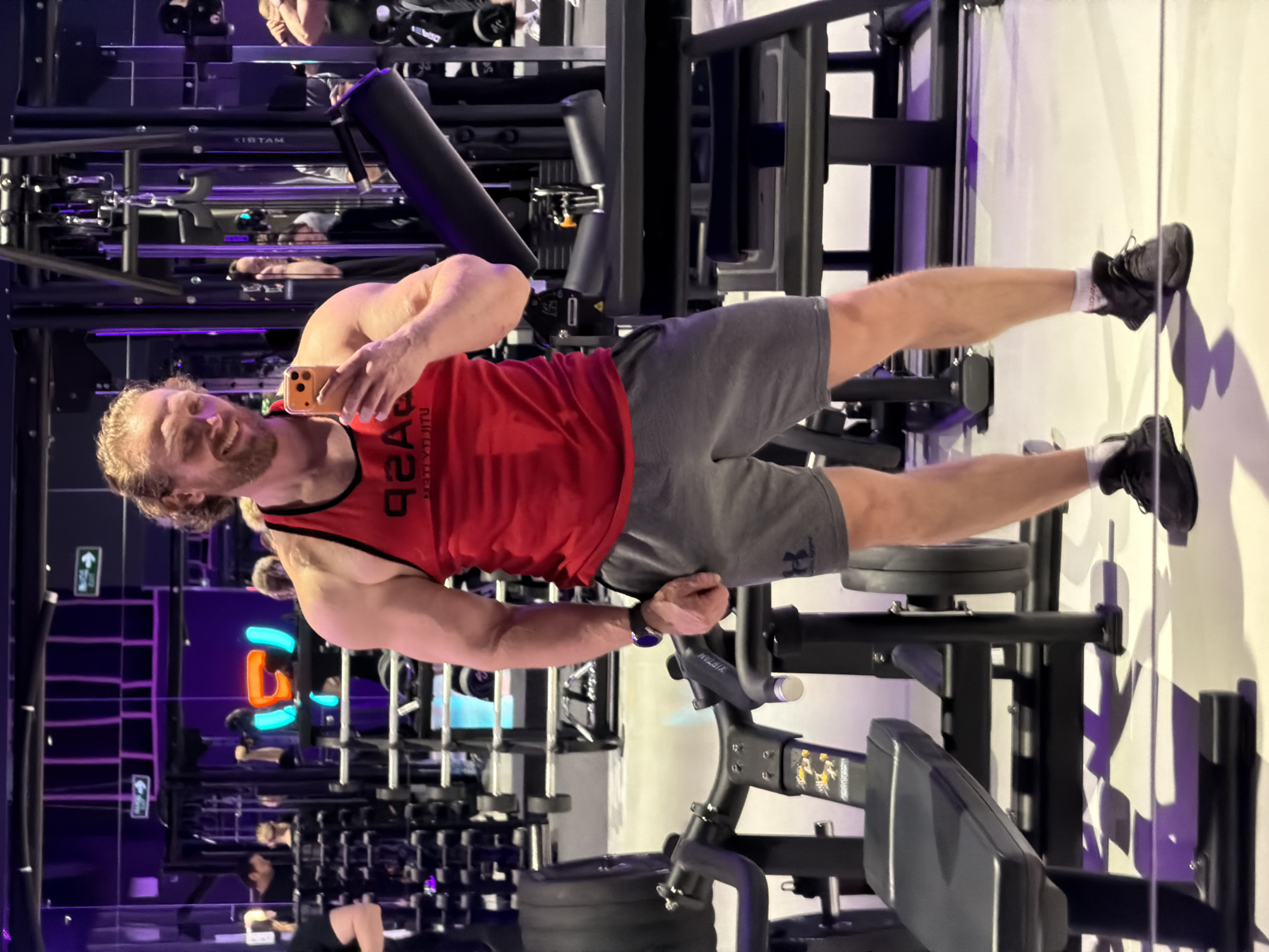 A man is taking a selfie in a gym. He is wearing a red tank top and gray shorts with black sneakers. The gym has various weight machines and free weights. There is a neon sign in the background. Other people are present in the gym, using the equipment. The gym appears to be modern with mirrored walls and a purple lighting scheme. There is no indication of Saint Petersburg, Russia in the image.