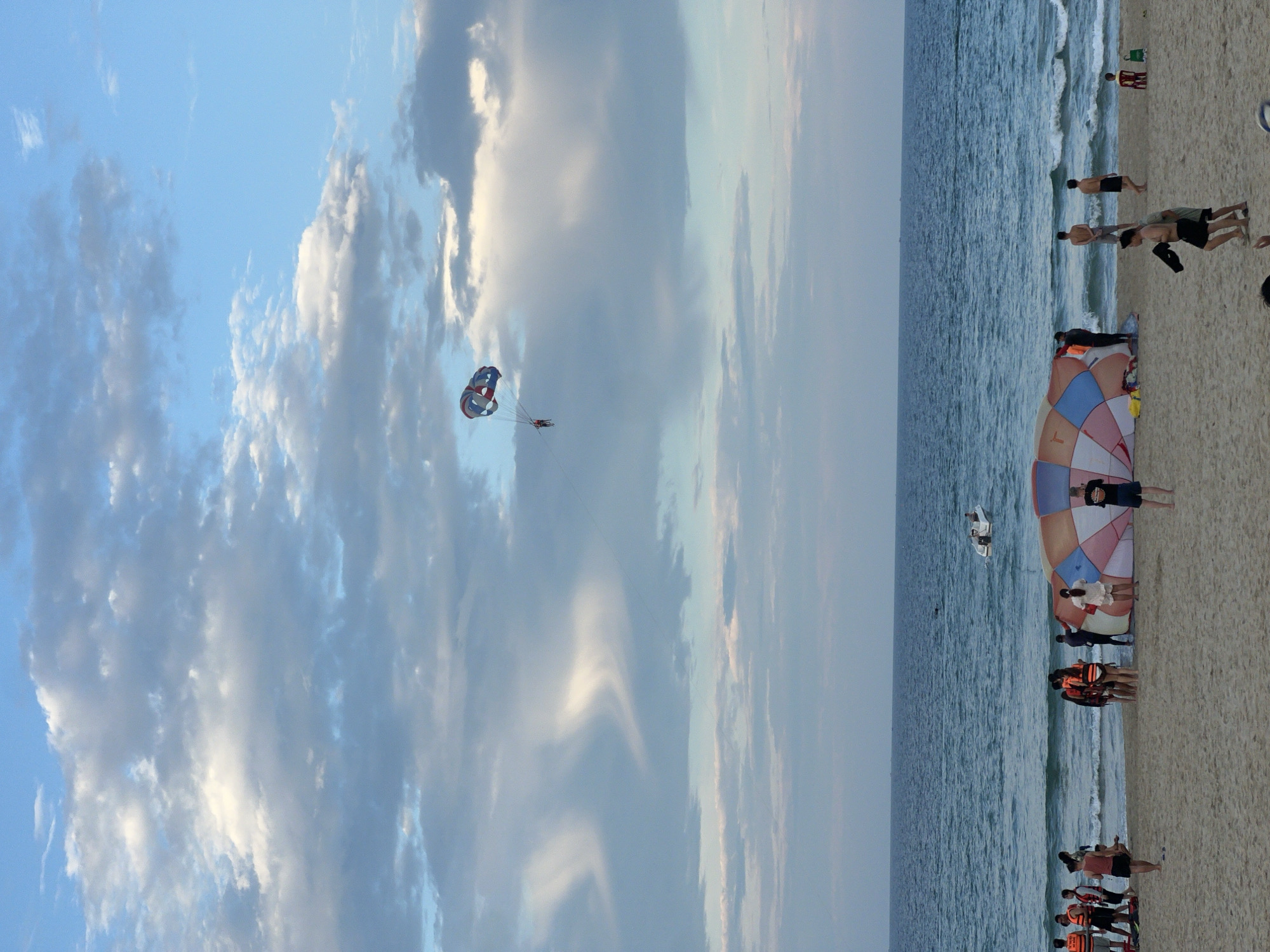 In the city of Unknown, Unknown, a beach scene unfolds under a sky filled with dynamic clouds. The ocean, a deep blue, stretches to the horizon, with a small white boat visible in the distance. On the sandy beach, a group of people are gathered around a large, partially deflated parachute, its colorful fabric spread out on the sand. Some individuals are wearing life vests, suggesting preparation for water activities. To the left, a group of people stand together, some wearing life vests, while others are observing the scene. In the sky, a person is parasailing, attached to a parachute, ascending into the vast expanse of the sky. The overall atmosphere is one of leisure and outdoor recreation.