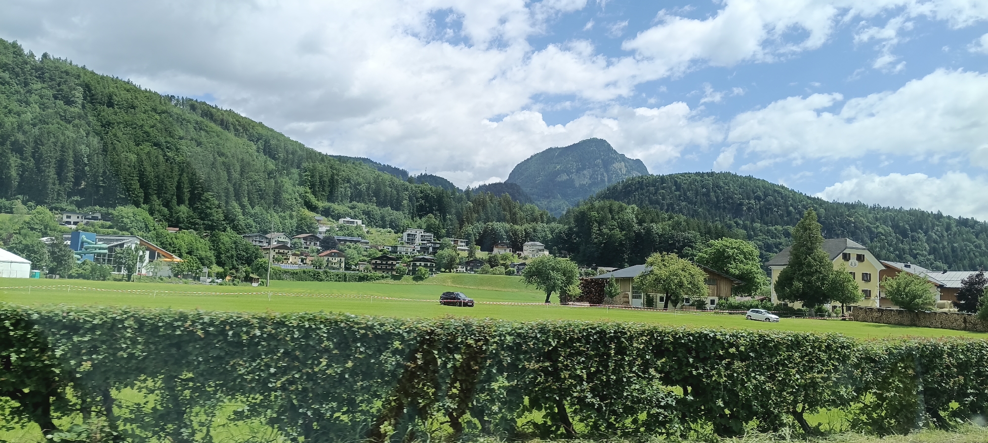 The image depicts a scenic view of the village of Torren, Austria, with a lush green field in the foreground bordered by a dense hedge. Beyond the field, a cluster of houses and buildings are nestled on a gentle slope, with a modern building featuring blue water slides prominent on the left. The background is dominated by a steep, tree-covered mountainside, with a large, rugged mountain peak rising above it. The sky is partly cloudy, with patches of blue sky visible. A dark-colored SUV is parked in the field, and a white car is visible further in the distance. The overall impression is one of a serene, picturesque rural landscape.