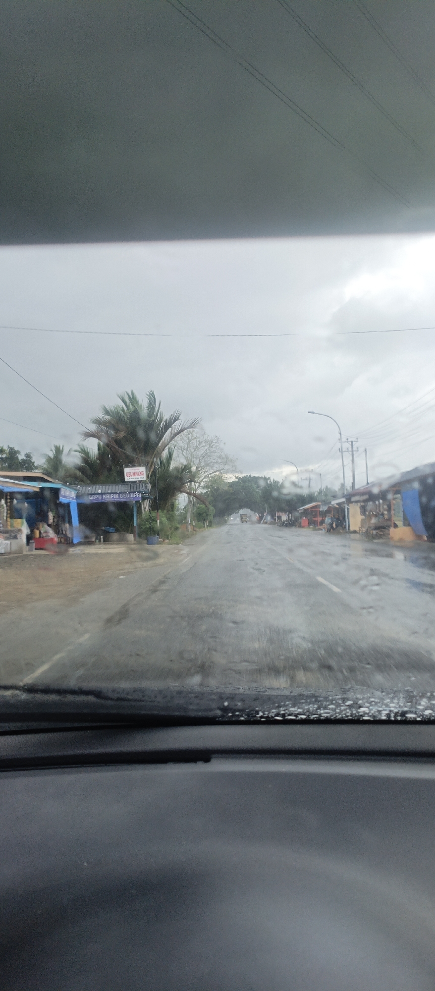 The image captures a road scene from the perspective of a vehicle's interior, looking out through a wet windshield. The weather appears overcast and grey, suggesting a recent rain or impending shower, with raindrops visible on the glass.
The asphalt road, visibly damp or wet, stretches into the distance, appearing to be a two-lane path with a white line marking the right edge in the midground. On the left side of the road, several small structures resembling shops or stalls are visible, some with blue awnings. Lush green trees, including palm fronds, stand behind these structures. A sign with red lettering reads "GELUMPANG", and below it, a blue sign displays "GAPU KRIPIK GELUMPANG", indicating a snack or chip shop.
To the right side of the road, more small buildings or stalls with varied colored tarps (red, yellow, blue) line the roadside. A utility pole with power lines and a street light fixture are also present. In the far distance, the road appears to pass under an archway formed by trees, with a vehicle, possibly a truck, barely discernible beyond. The foreground shows the car's dashboard and the lower part of the windshield, emphasizing the indoor perspective. The scene suggests a rural or semi-urban area in Glee Gogo, Indonesia, likely during daytime given the ambient light, despite the heavy clouds. No people are clearly visible engaged in specific activities, though the presence of shops implies commercial activity.