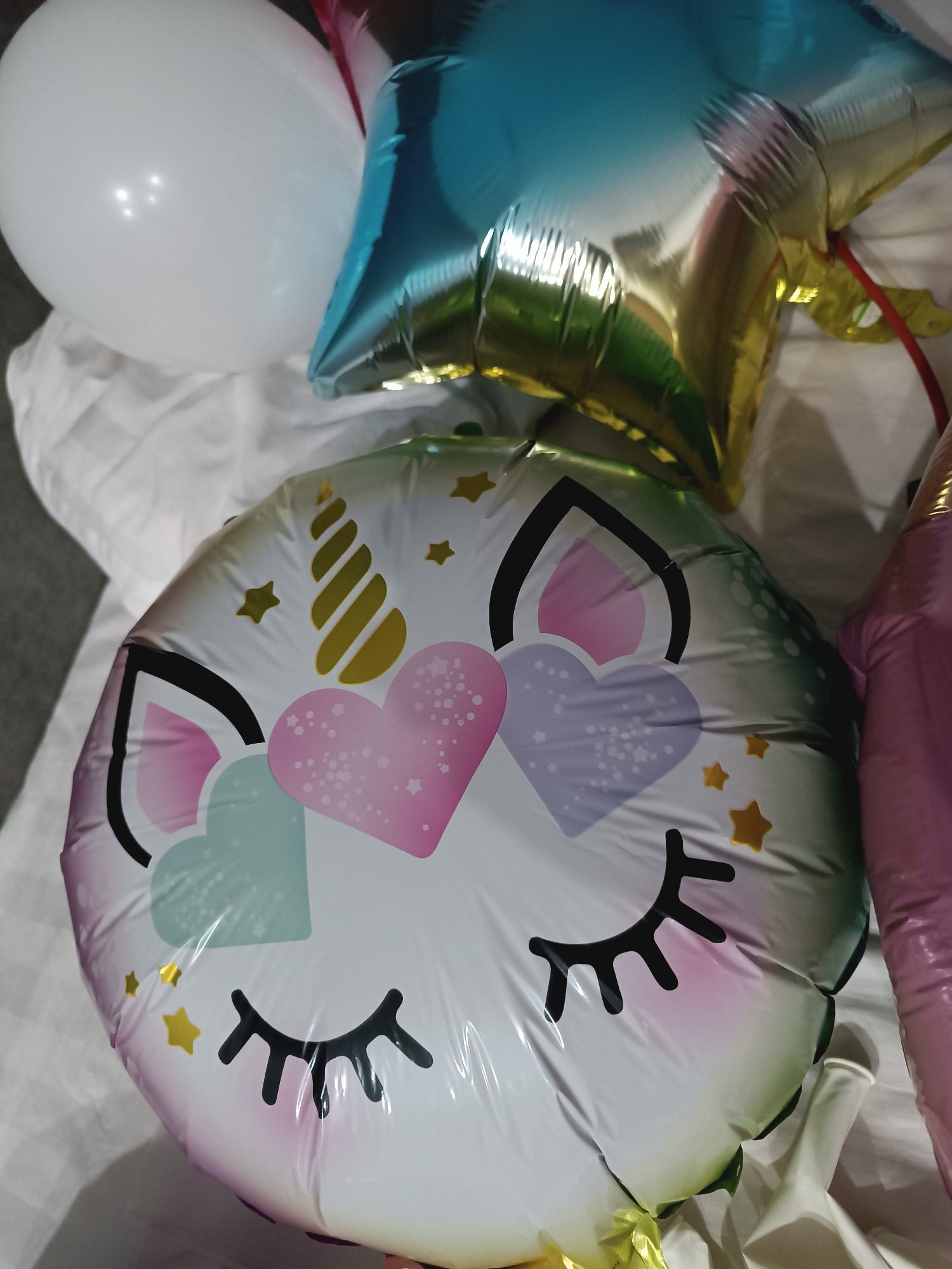 The scene depicts a close-up, top-down view of multiple balloons resting on a white, textured fabric surface, identified as bedding, within an indoor domestic environment in Unknown, Unknown. The central object is a deflated or partially inflated circular foil balloon featuring a cartoon unicorn face design. This balloon displays closed black eyelashes, a golden spiral horn, pink ears, and three heart-shaped designs in pink, light blue, and lavender, along with scattered golden stars on a white background. To the upper left, a fully inflated, smooth white latex balloon is visible. To the upper right, partially obscured, is a multi-colored foil star-shaped balloon, predominantly blue and gold, with a red ribbon attached. A segment of another pink foil object, likely a balloon, is visible along the right edge of the frame. No individuals or active interactions are present.