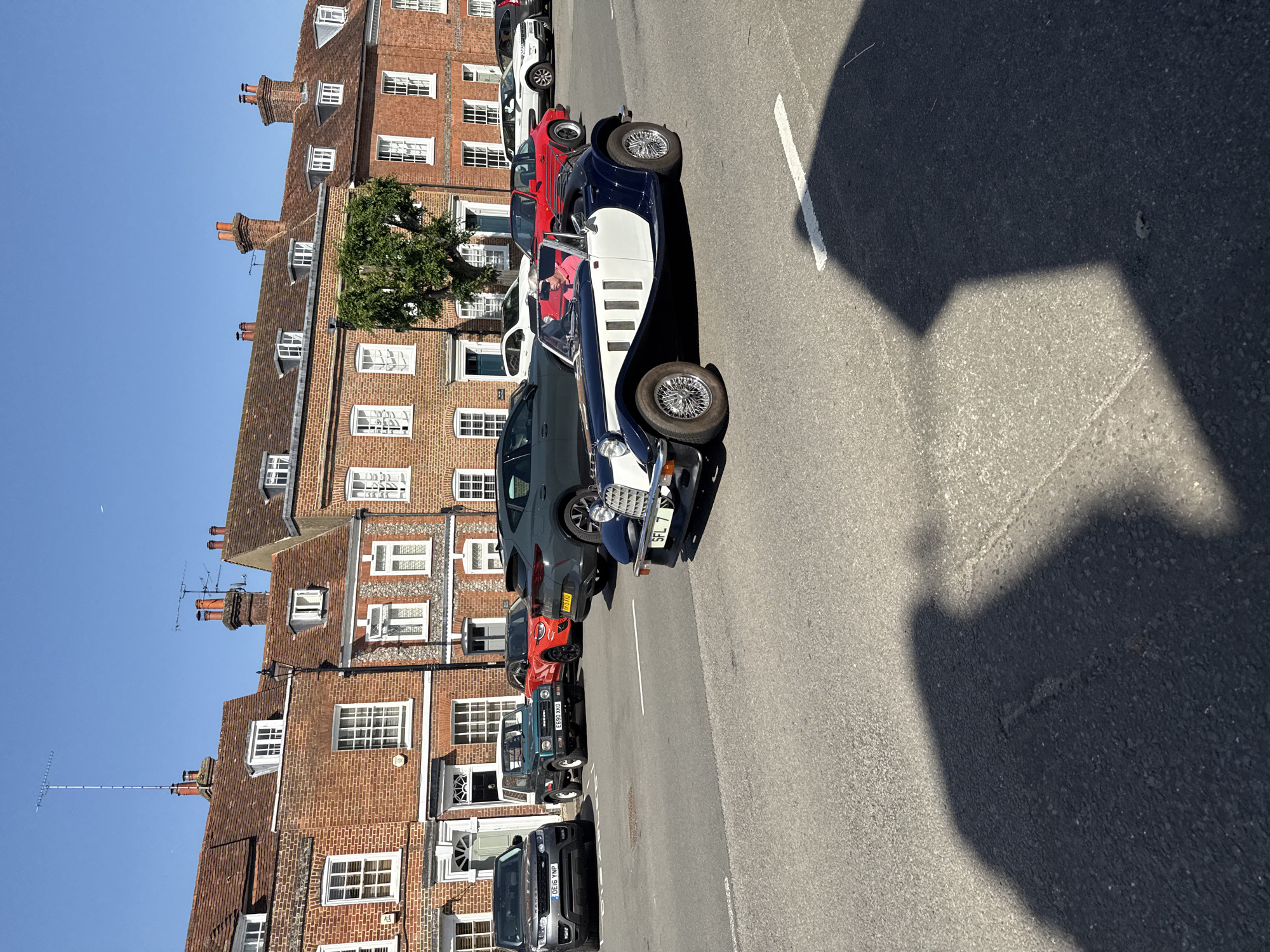 The image depicts a street in Amersham, United Kingdom, characterized by brick buildings with multiple windows and gabled roofs, lined with parked vehicles. The foreground is dominated by a dark blue and white vintage-style car with wire wheels. Behind it are parked several other cars of various colors and models, including a red car and a dark-colored SUV. A light-colored line marking is visible on the road surface. A tree is visible in front of one of the buildings. The sky is clear and blue. The license plate of the vintage car reads "SFL 7." The license plate of a dark gray car reads "M15 HW." The license plate of a black car reads "ESS9 XKO." The license plate of a black car reads "GE16 YWP." A person is seen inside the vintage car. Shadows are cast on the road from the buildings and objects.