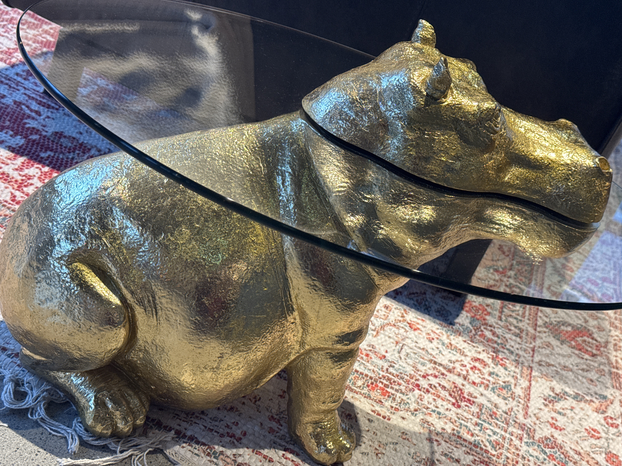 A golden hippopotamus sculpture is positioned on a patterned rug. The sculpture is textured and appears to be made of metal with a gold finish. A black, curved glass tabletop is situated on top of and partially over the hippopotamus, casting shadows and reflections. The rug beneath has a red, white, and blue pattern. The background is dark, suggesting an indoor environment, possibly a living room. The image is taken from a high-angle perspective. There are no people present in the scene.
