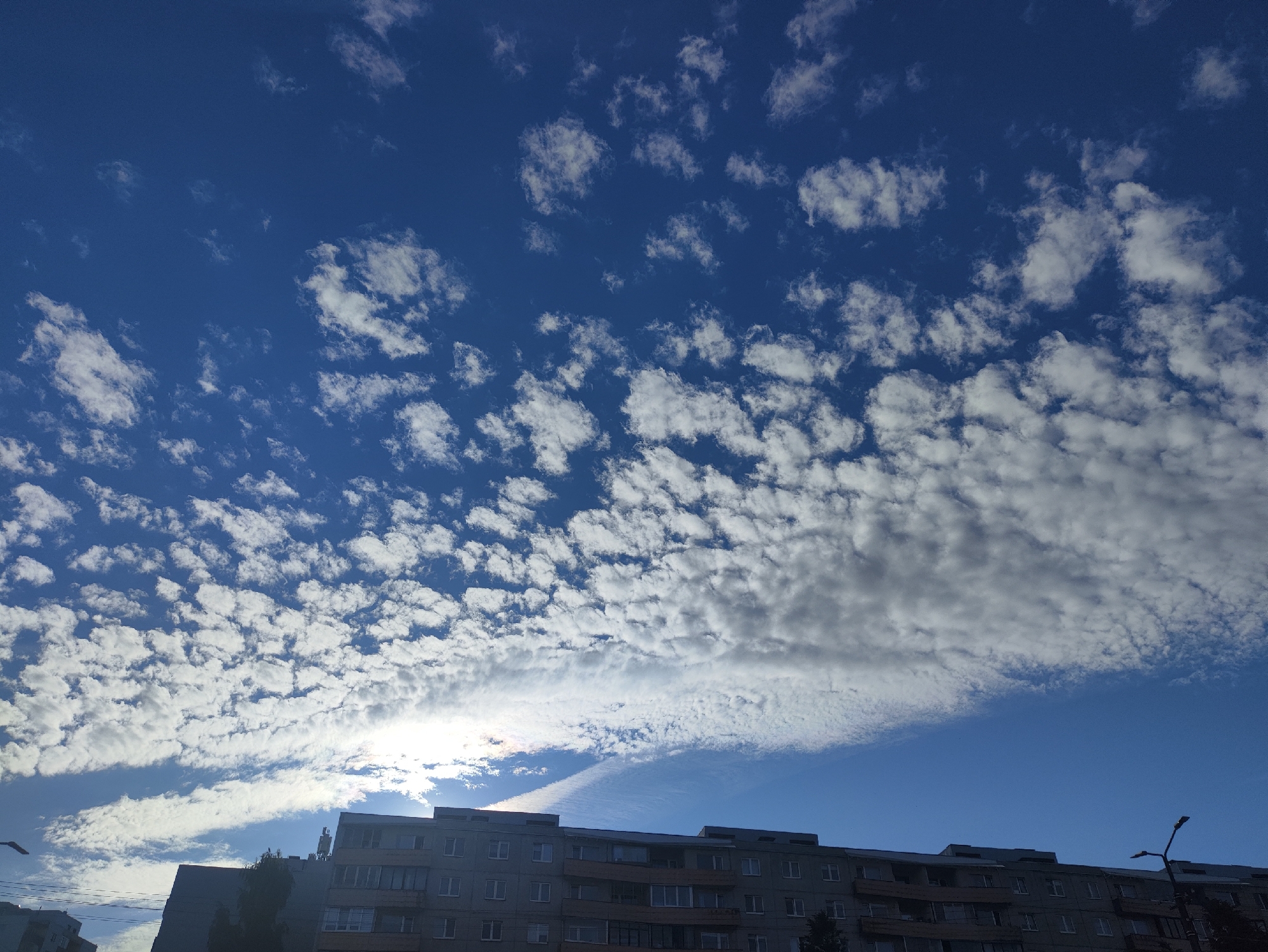 This is a horizontal, eye-level shot of a bright, sunny day with a clear blue sky filled with scattered clouds. The clouds are a mixture of fluffy white cumulus clouds and thinner, wispy cirrus clouds forming a band across the sky. The sun is visible behind the clouds on the left side of the frame, casting a bright glow.
In the foreground, the tops of several apartment buildings are visible. The buildings are multi-story structures with a grid of windows and balconies. The architecture suggests a European setting, consistent with the provided location context of Tallinn, Estonia. A street lamp is visible on the right side of the frame, silhouetted against the sky.
The overall impression is one of a pleasant, albeit partly cloudy, day. The bright sunlight and blue sky suggest it is daytime, likely late morning or early afternoon. There is no discernible activity or event taking place. No visible text is present in the image.