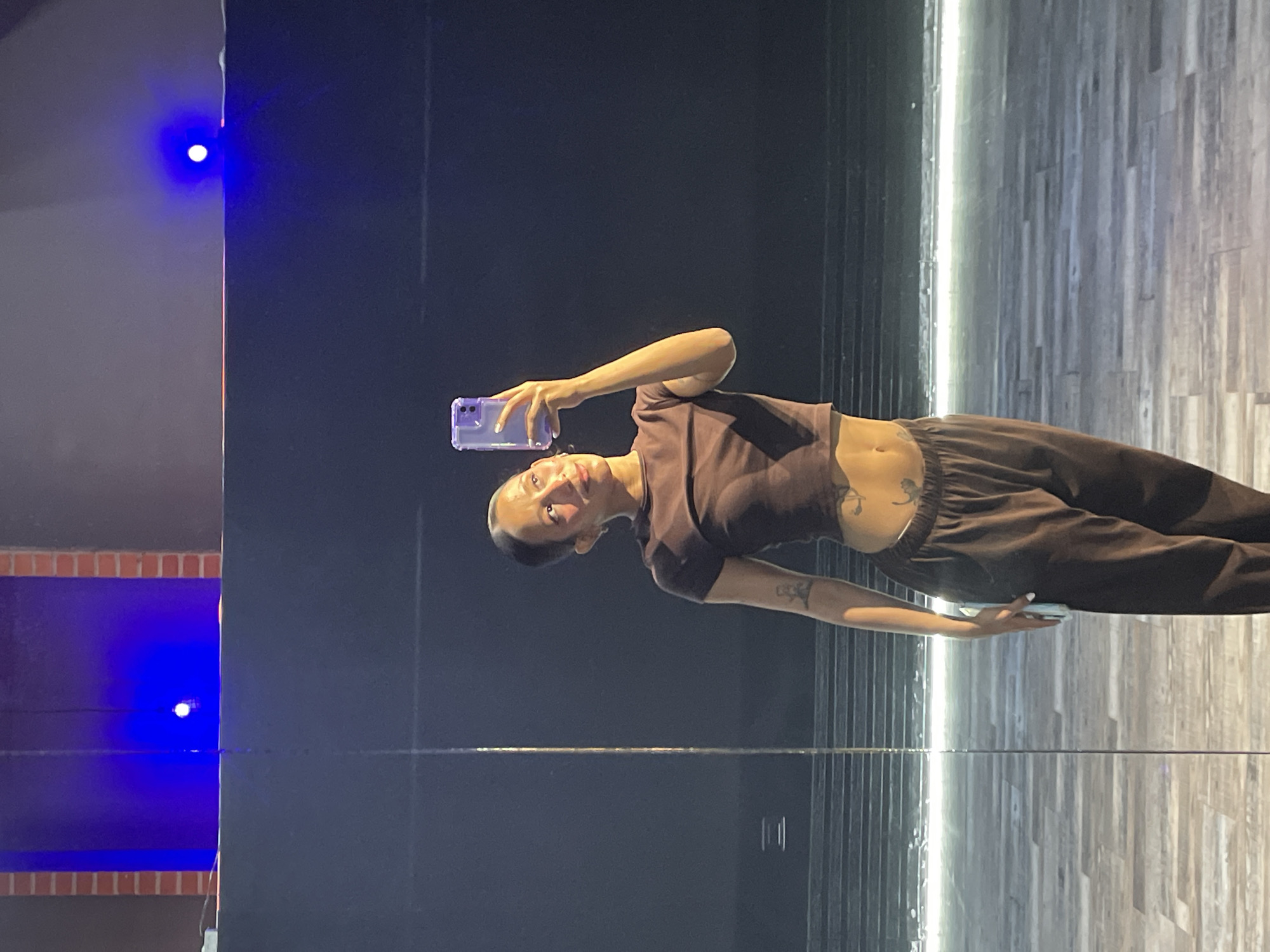 A young woman with dark hair pulled back into a bun is taking a selfie with a purple-cased smartphone in a dimly lit studio. She is wearing a brown crop top and matching brown sweatpants. The woman has tattoos on her stomach and arm. The background of the image shows a large dark wall, a reflective floor, and two bright blue lights, one on the left and one on the right. The overall setting suggests an artistic or performance space. The woman is positioned in the center of the frame, looking towards her phone screen.