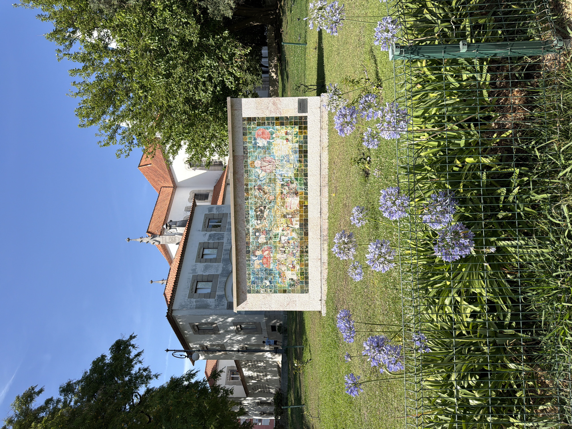 The photograph shows a tiled mural on a building in Lisbon, Portugal. The mural depicts a scene with people dressed in historical clothing, engaged in various activities. The mural is set against a backdrop of a building with a red-tiled roof and white walls. In the foreground, there are purple flowers growing in a garden, separated by a green metal fence. The sky is clear and blue. A person is visible in the background, standing near the building. The overall setting suggests a public space, possibly a park or a historical site within the city.