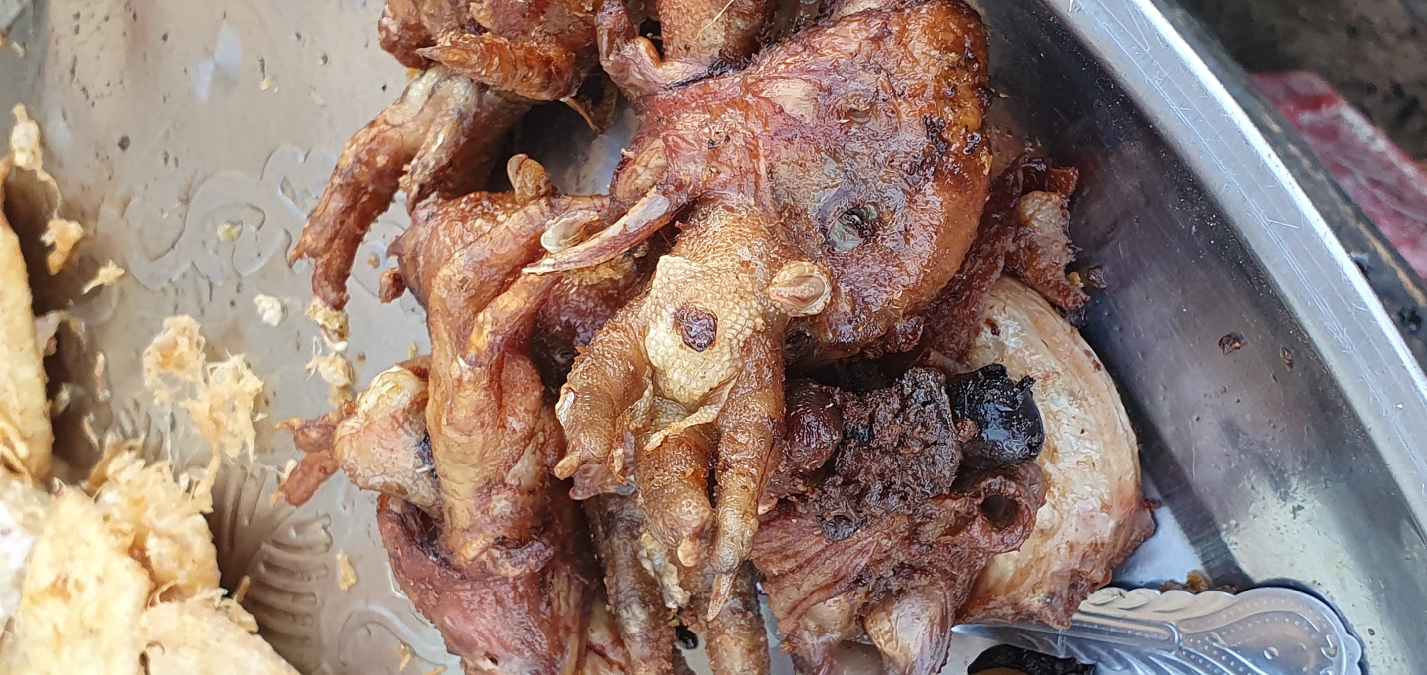 A close-up shot shows a pile of deep-fried chicken feet on a metal tray. The chicken feet are golden brown and appear crispy. Some of them have visible skin and bone structure. The tray is slightly reflective and shows some scratches and smudges. The background is out of focus but suggests an outdoor setting with natural light. The overall presentation is casual and suggests a street food vendor or a home-cooked meal. The image is likely taken in Bukuru, Nigeria, given the context provided.