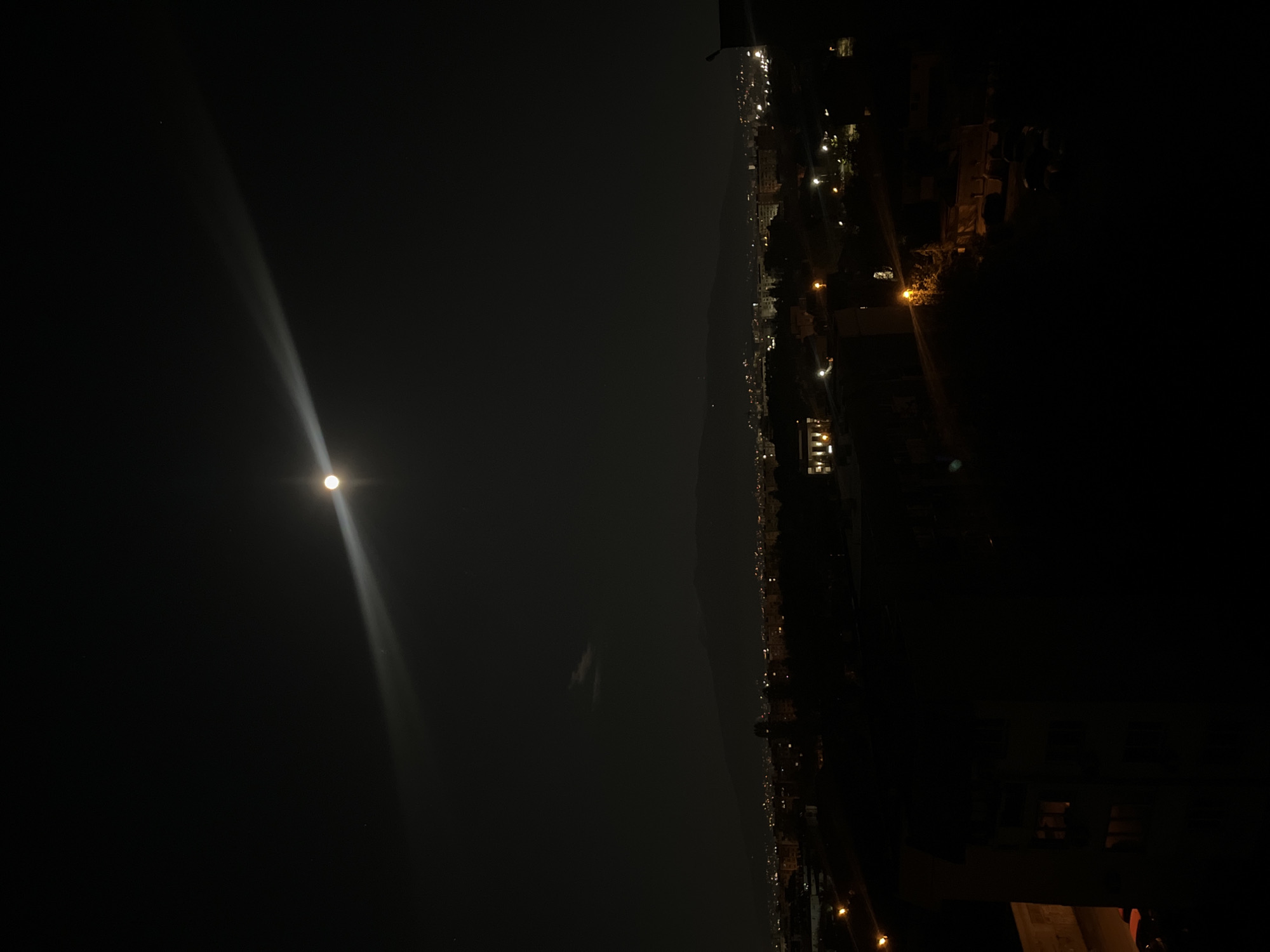 The image shows a night view over an urban area. A bright moon is visible on the left side of the frame, with a beam of light extending downwards. On the right side, a city line is illuminated with various lights, indicating buildings and infrastructure. A dark mountain range is discernible between the moon and the cityscape. Based on the general appearance of the urban area, the presence of a mountain range, and the style of lighting, it is possible that the scene is in Sofia, Bulgaria.