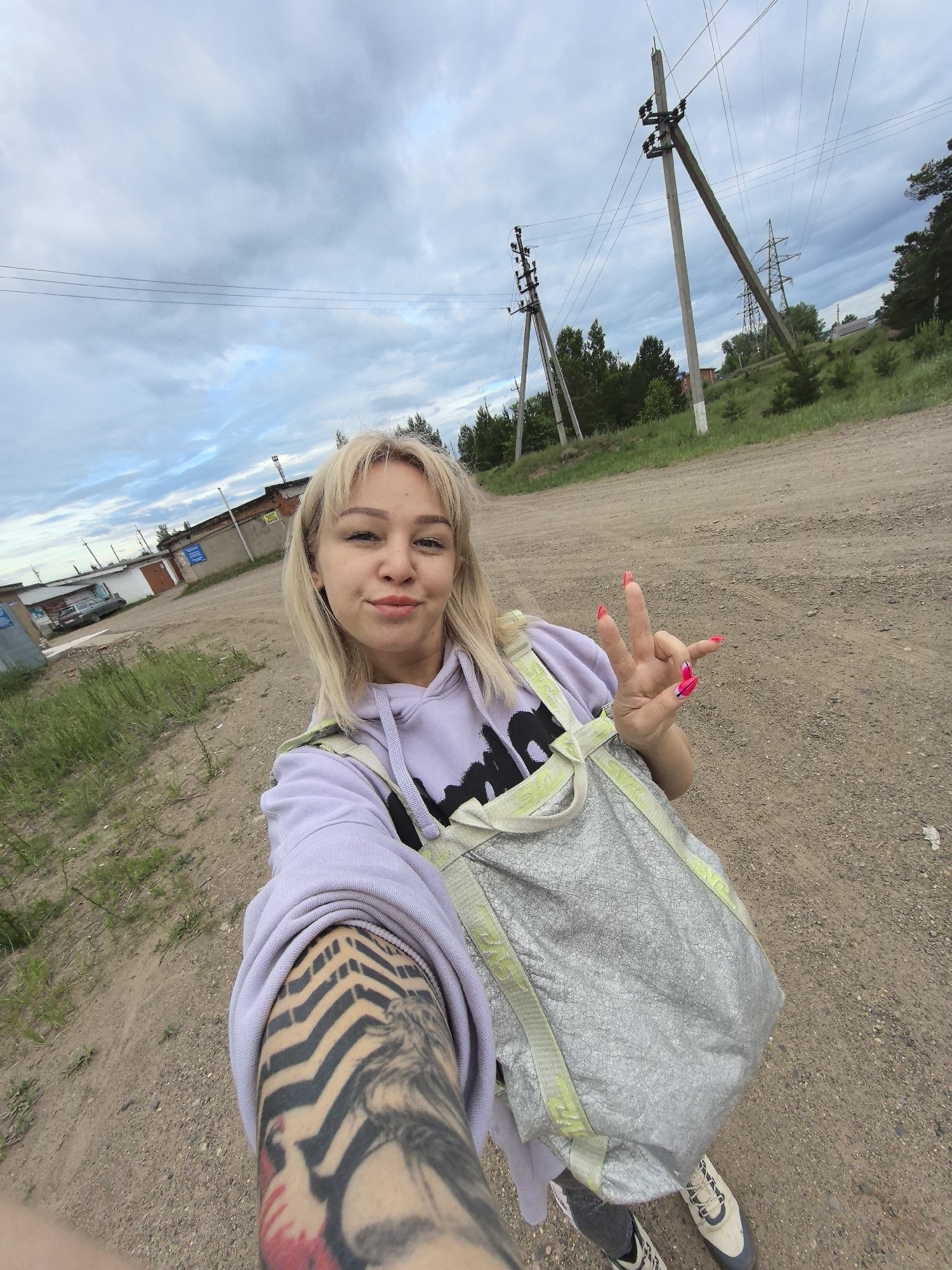 A blonde female individual is centered in a close-up selfie, captured outdoors on an unpaved dirt road in Noviy, Russia. She is wearing a light purple hooded sweatshirt, a textured grey tote bag with light green straps, and white and grey sneakers. Tattoos consisting of black chevron patterns and other dark designs, including some red elements, are visible on her left arm. Her right hand is raised, forming a three-finger gesture (index, middle, and ring fingers extended), with her fingernails painted a vibrant pink with darker tips. Her facial expression displays a slight, closed-lip smile. The environment features a dirt track with sparse grass. To the left, several single-story, possibly industrial or garage-like buildings are visible, with a dark grey sedan parked near one. To the right and in the background, a landscape of trees and green foliage extends under an overcast sky. Multiple utility poles and overhead power lines are prominent, with one pole leaning noticeably. A large metal electricity transmission tower is also present in the distant background.
