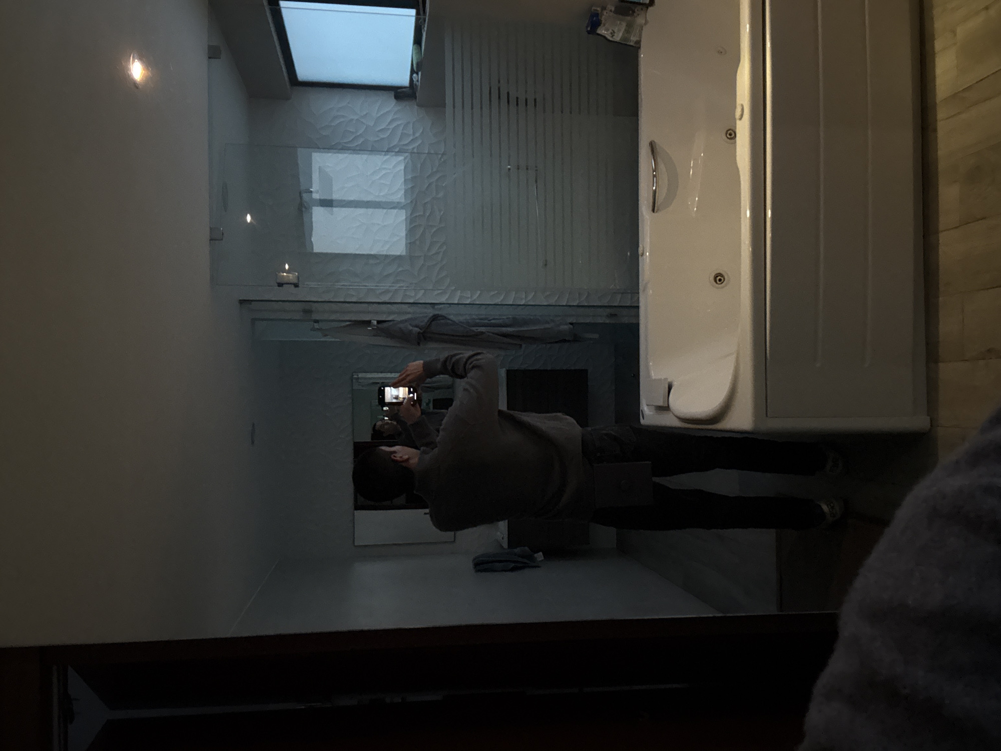 This image depicts the interior of a modern bathroom, presumably located in San Bernardino Tlaxcalancingo, Mexico. A male individual, viewed from behind, stands in the center-left, wearing a gray long-sleeved shirt, dark pants, and what appears to be a dark pouch on his right hip. He is holding a smartphone horizontally with both hands, aiming it towards a large mirror positioned above a dark double vanity. His reflection is visible in the mirror, showing him in the act of taking a self-portrait.
To the right of the subject and occupying the foreground is a large white built-in bathtub with a silver handle and multiple jets. Behind the bathtub is a glass-enclosed shower stall featuring frosted horizontal privacy strips. A textured white wall with a wave pattern is visible behind the shower enclosure. A flat-screen television is mounted on the wall to the far right. The floor is covered with light-toned wood-look planks. The bathroom is dimly lit, with recessed lighting visible in the ceiling and an illuminated smartphone screen. A dark gray towel hangs on the wall to the left of the vanity, and various toiletries are visible on the edge of the tub.
