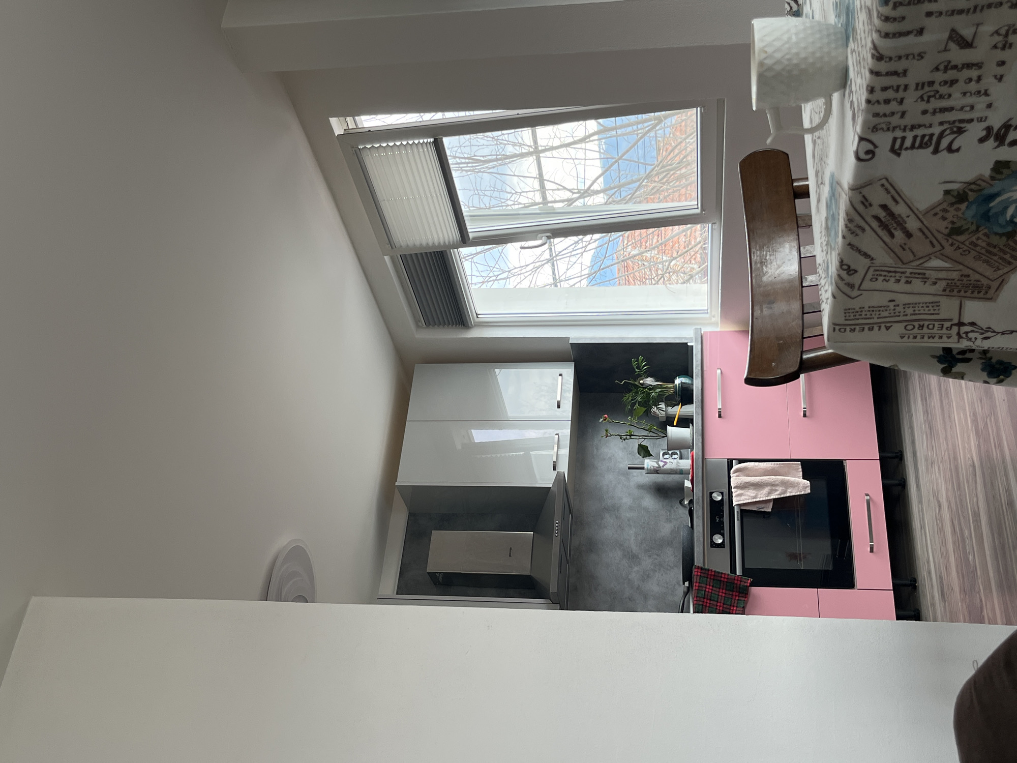 A high-angle shot shows a kitchen area in an interior space with a slanted roof. The kitchen features white cabinets, a modern range hood, and pink kitchen units with a built-in oven. A window in the slanted roof allows daylight to illuminate the scene. A dining area with a table covered with a patterned tablecloth and a white mug on the table is visible. The presence of indoor plants adds a touch of nature to the space. The location in the city of Karavelovo, Bulgaria is not discernible from the image.