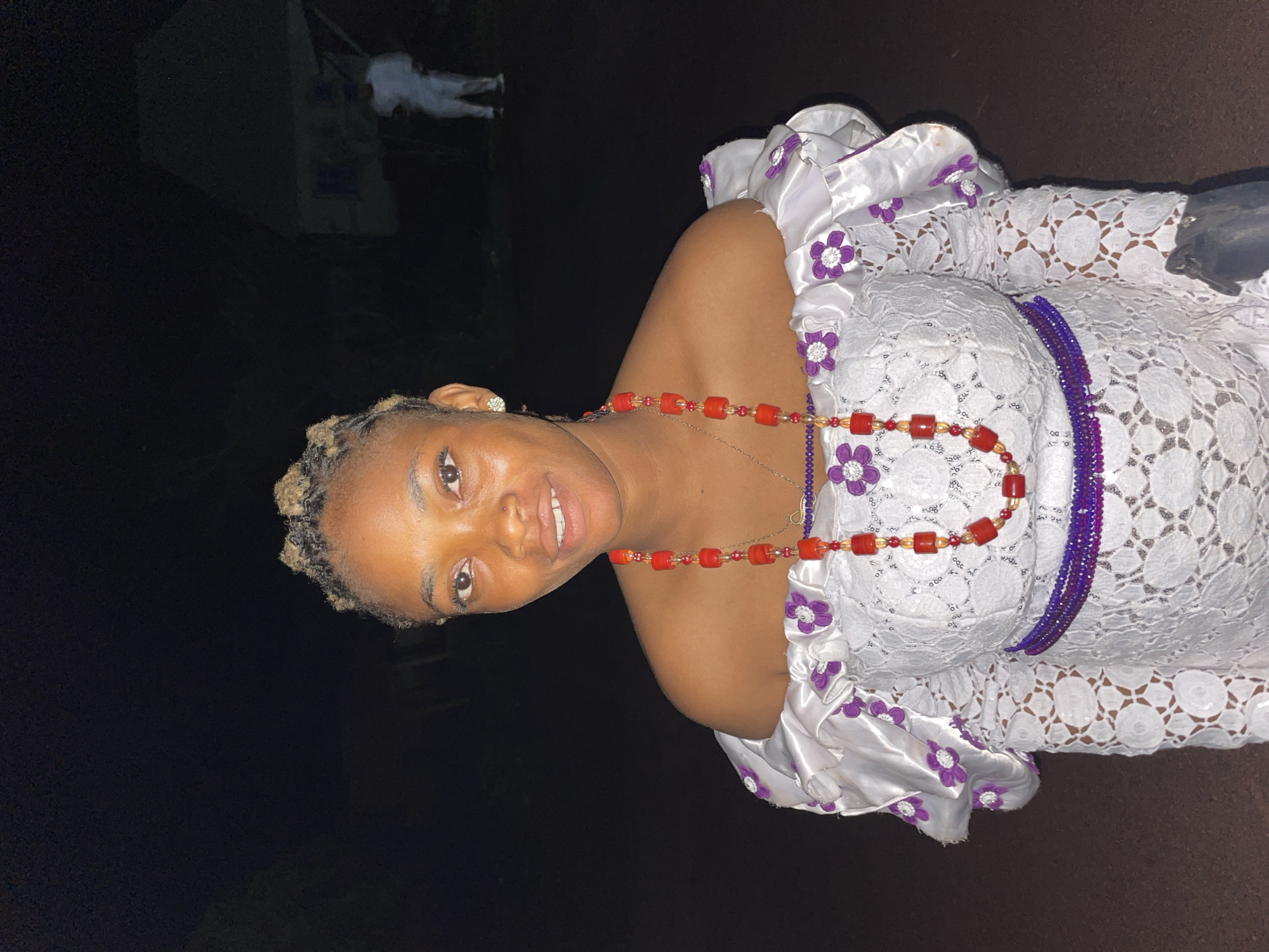 A woman is standing outdoors at night. She has light skin and short, light-colored hair. She is wearing a white, off-the-shoulder dress with purple flower embellishments and a lace overlay. She is also wearing a red beaded necklace. A purple beaded belt or necklace is visible on her waist. The background is dark, with a faint image of a person in white clothing in the distance. The location may be Ogbe, Nigeria.