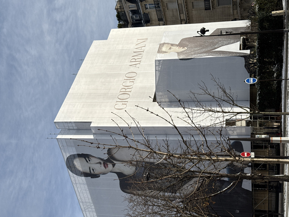 The scene depicts the Giorgio Armani flagship store located at 18 Avenue Montaigne in Paris, France, currently encased in white protective sheeting due to renovation or construction. The sheeting functions as a large advertising canvas. On the left facade, a prominent banner features a female model of East Asian descent, styled with dark hair, a dark garment, and red lipstick, looking towards the viewer. On the right facade, the "GIORGIO ARMANI" brand name is displayed in large, light-colored lettering, positioned above a vertical banner featuring a male model with light hair, dressed in a dark, textured top, also looking towards the viewer.
In the foreground, several leafless deciduous trees partially obscure the lower sections of the building. The ground below the trees exhibits a thin, patchy layer of snow or frost, suggesting cold weather conditions. A red circular "No Entry" traffic sign and a blue circular "Mandatory Ahead" sign are visible at street level. To the far right, an ornate, multi-story Parisian building with balconies and a dark roof is visible adjacent to the Armani structure. The sky is partly cloudy with visible blue areas, indicating natural daylight illumination.