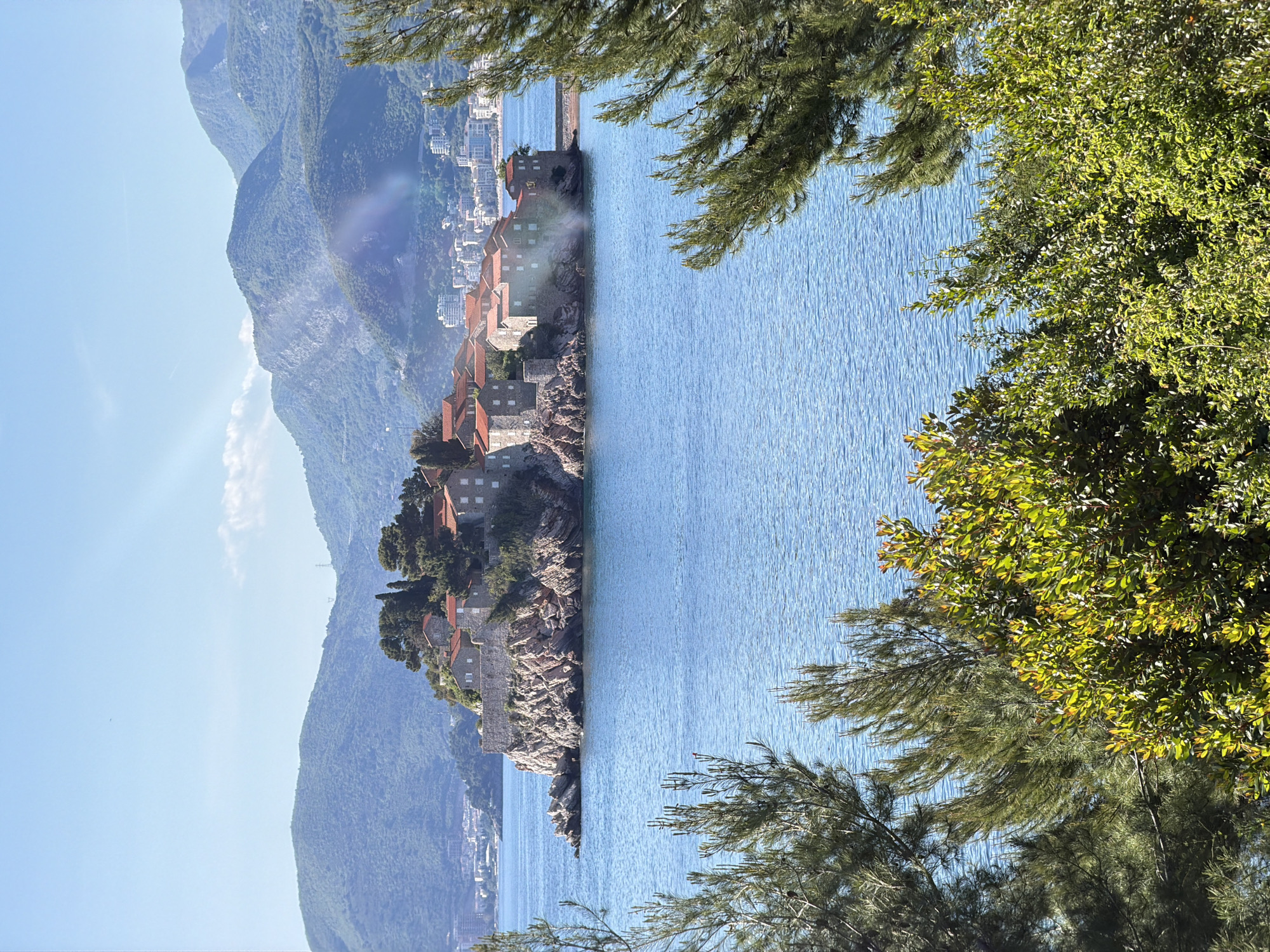 The image captures Sveti Stefan, Montenegro, showcasing the island resort with its terracotta-roofed buildings. The Adriatic Sea surrounds the island. The backdrop features mountains and a clear blue sky. Foliage in the foreground partially obscures the view.