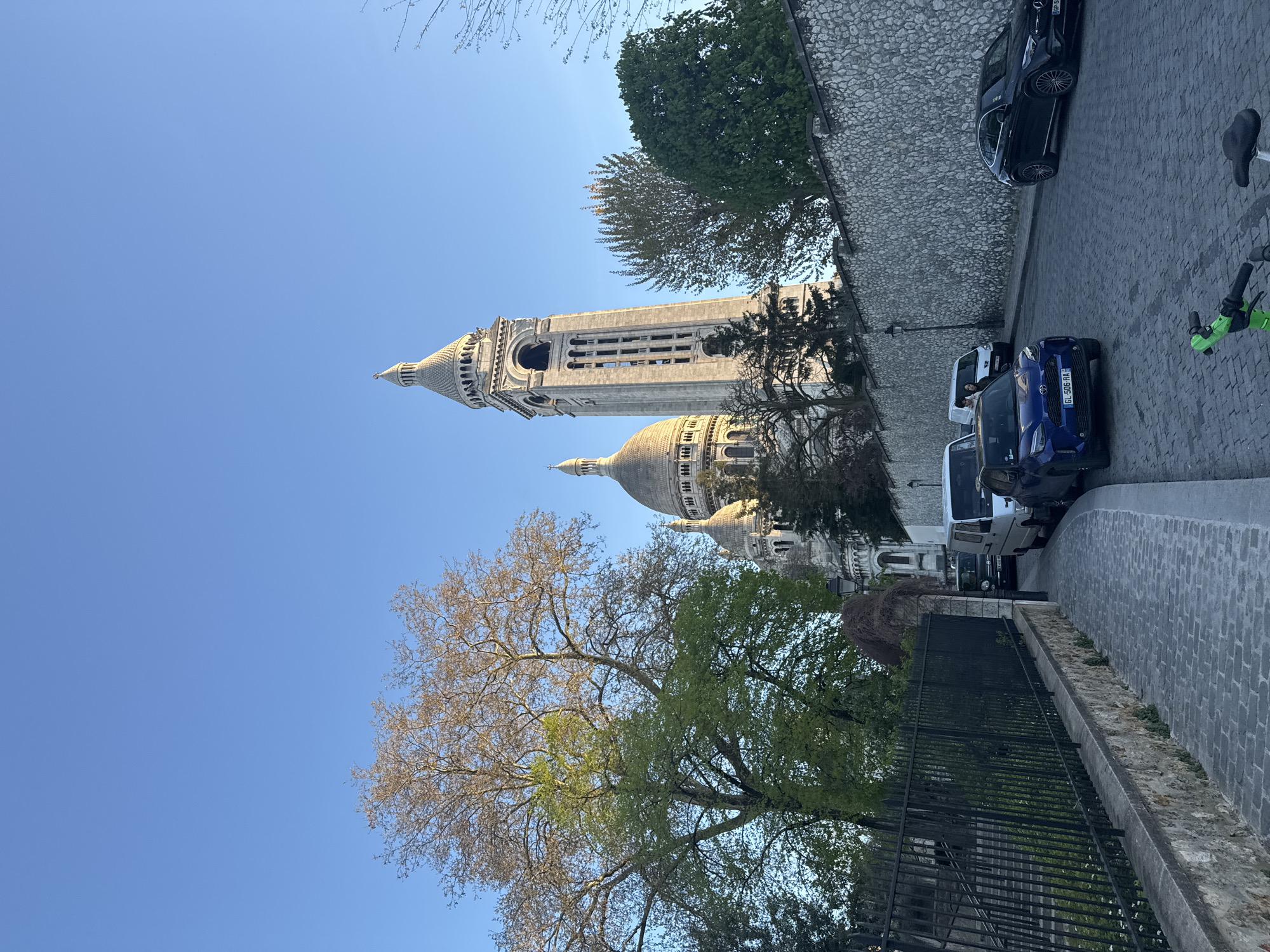 The image shows the Sacré-Cœur Basilica in Paris, France, viewed from a street level perspective. A tall, ornate tower and the basilica's dome are visible. The street is paved with cobblestones and lined with parked cars, including a blue vehicle with license plate "GL-506-RA." A black fence and trees frame the view. The sky is clear blue.