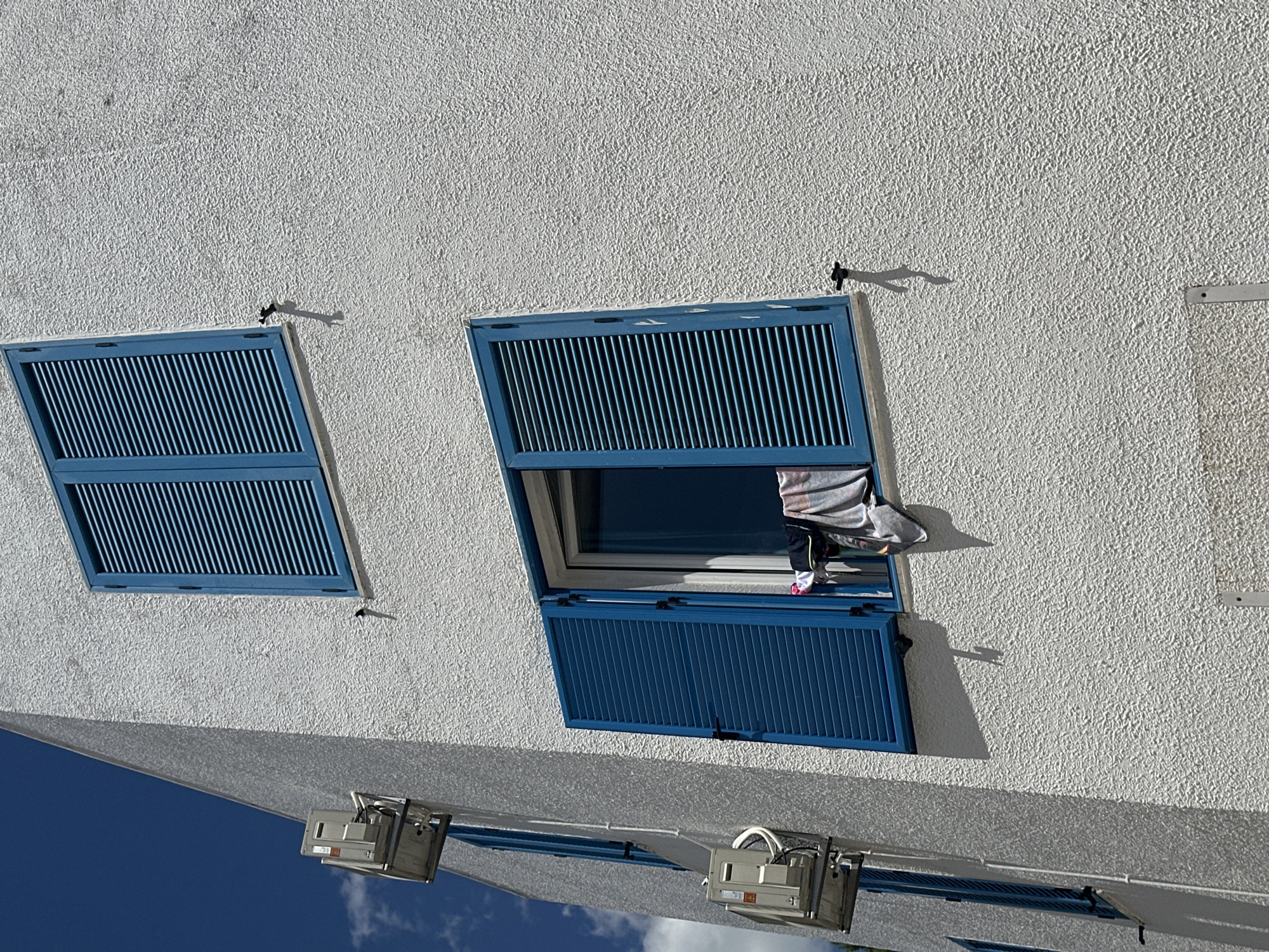 A white stucco building with blue shutters is seen under a bright blue sky with sparse clouds. The building has at least two windows with blue shutters, one of which is open. On the open window, laundry is hung to dry, including a white towel and darker clothing. There are also two air conditioning units mounted on the side of the building. The building appears to be located in Budva, Montenegro. The scene is observed from a low angle looking upwards.
