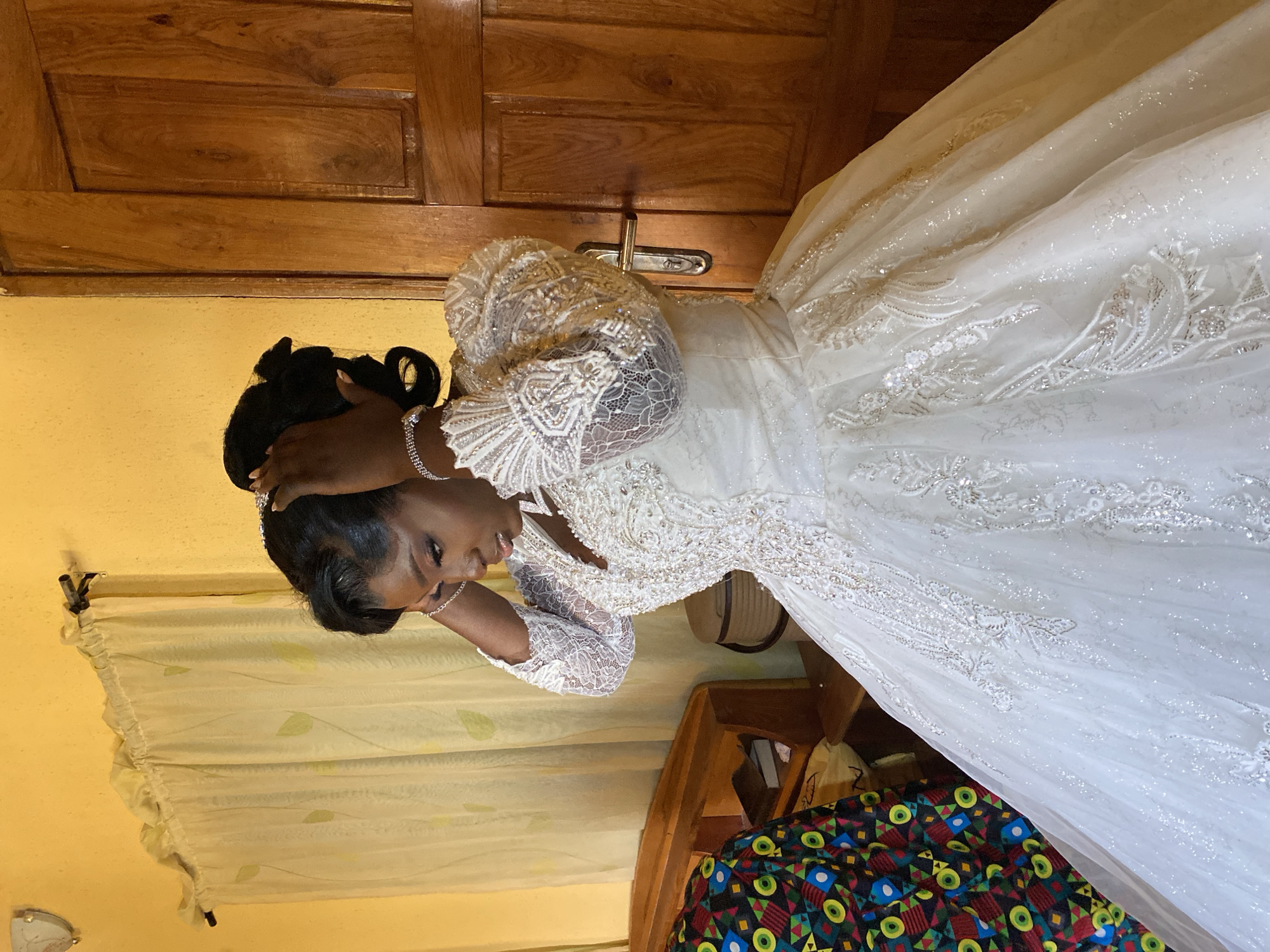 A woman stands in a room, dressed in a white wedding gown. She is holding her hair, likely adjusting her hairstyle. Behind her is a wooden door with a metal handle and a curtained window. The photograph was likely taken in Vom, Nigeria.