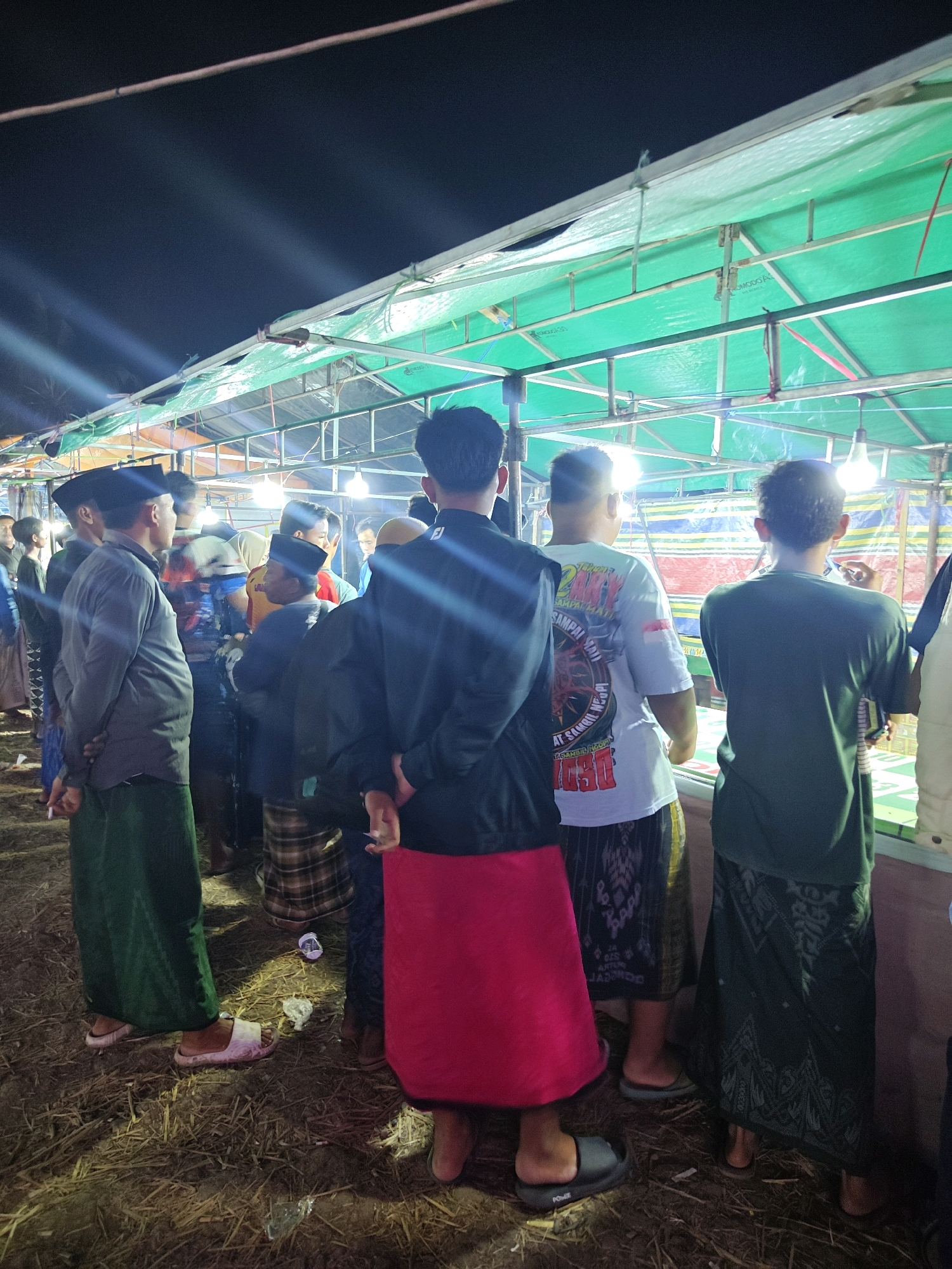 This is a nighttime, eye-level, medium shot of a group of men standing under a green tarpaulin canopy supported by a metal frame. The scene appears to be outdoors, with dry grass and some litter on the ground. Several bare lightbulbs are strung overhead, illuminating the area. Most of the men are wearing traditional sarongs, and some are wearing skullcaps.
In the foreground, a man on the left wears a grey long-sleeved shirt and a green sarong. To his right, another man wears a black jacket and a bright pink sarong. Further back, a man in a white t-shirt with graphics stands next to a man in a dark green t-shirt and a patterned sarong. More individuals are visible in the background, some facing away from the camera. One man in the middle is smoking. The overall atmosphere suggests a casual gathering or event in Suling Wetan, Indonesia, possibly a night market or a religious ceremony.
