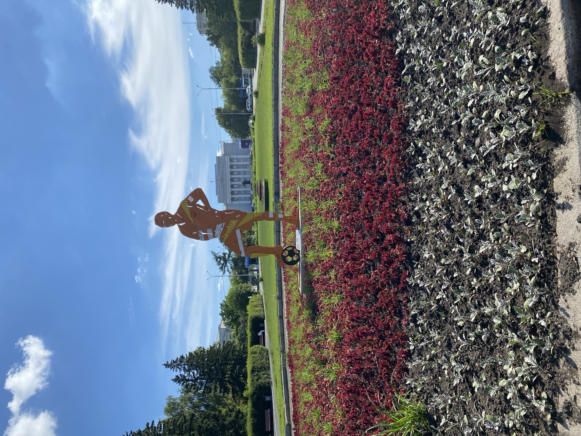 The image shows a metal sculpture of a man with a soccer ball at his feet, positioned in the center of a flower bed. The sculpture is orange and depicts a stylized human figure. The flower bed features a band of red plants and a band of silver-green plants at the bottom. In the background, there is a large building with columns, surrounded by green trees and a blue sky with scattered clouds. The scene appears to be a public space, possibly a park or a plaza, in the city of Yekaterinburg, Russia. The sculpture is located near the Yekaterinburg City Administration building.