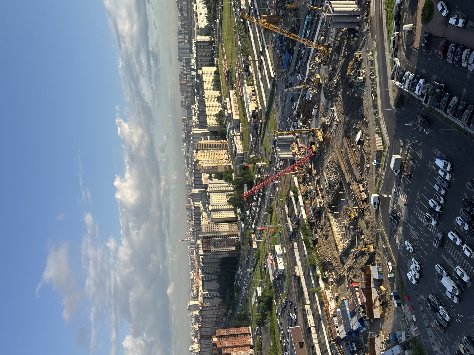 The image depicts an aerial view of an active construction site in Saint Petersburg, Russia, likely in a newly developing or redeveloping urban area. The foreground is dominated by the construction site itself, with heavy machinery such as red and yellow cranes, excavators, and drilling equipment actively engaged. Piles of dirt, concrete structures, rebar, and temporary buildings are visible. In the middle ground, there is a large parking lot filled with various cars, including sedans, SUVs, and a white van, indicating a populated area adjacent to the construction. A road with a moderate amount of traffic can be seen bordering the parking lot and construction site. Further in the background, a dense cityscape of residential buildings, characterized by multi-story apartment blocks, stretches across the horizon under a partly cloudy blue sky. The architecture suggests a typical Soviet-era or post-Soviet urban development. The lighting indicates it is either early morning or late afternoon, with the sun casting long shadows. The overall scene captures the dynamic process of urban development and infrastructure expansion within a city.
