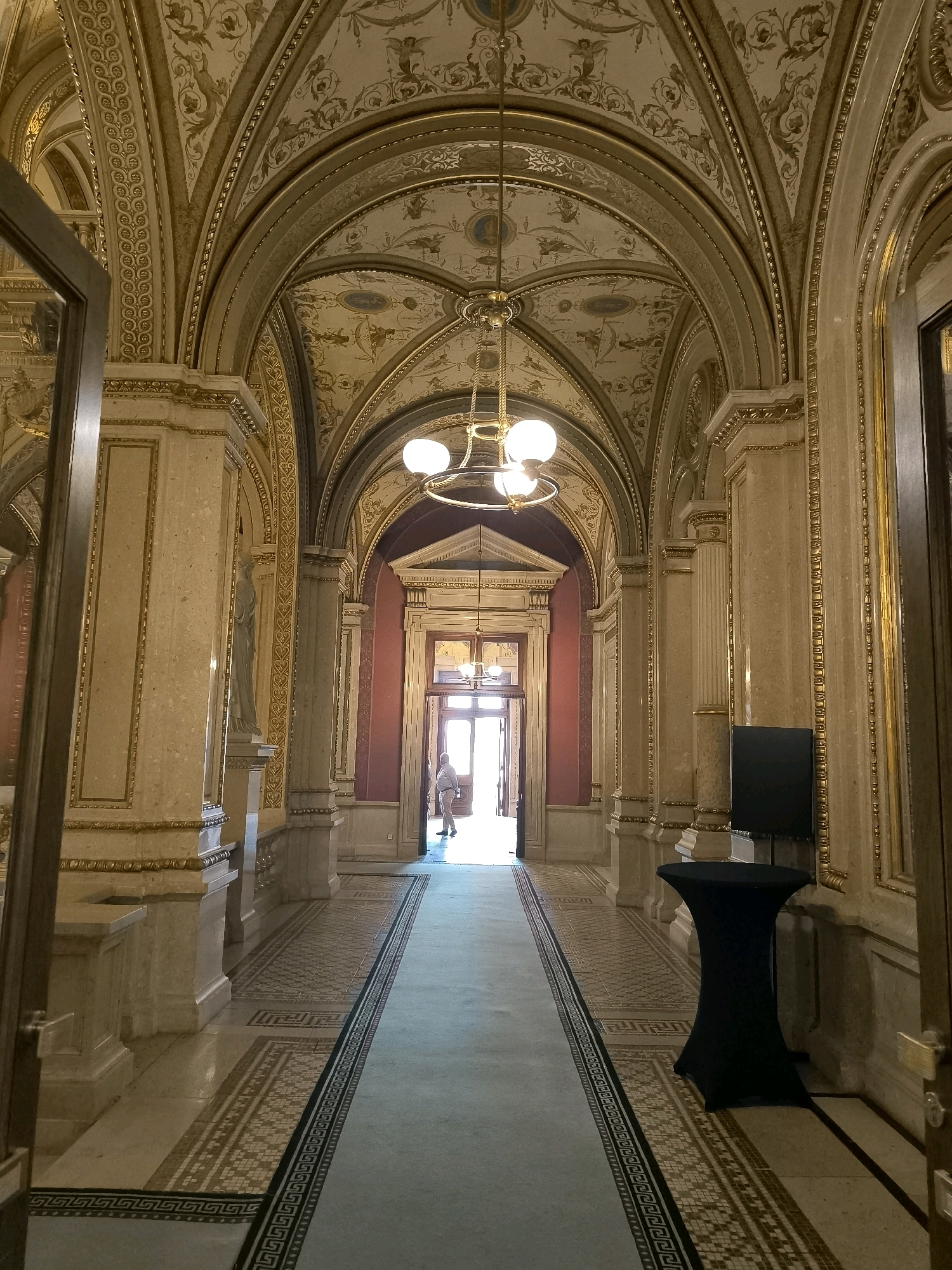 This is a low-angle, full shot photograph of a grand, ornate hallway with a high, arched ceiling, likely within a historic building in Vienna, Austria. The hallway is illuminated by a large, decorative chandelier and natural light filtering in from a doorway at the far end. The ceiling is adorned with intricate gold and cream-colored frescoes depicting mythological figures and decorative patterns.
The walls are lined with tall, marble-clad columns with gold detailing and ornate capitals. Between the columns, there are decorative panels and a life-sized statue of a robed figure. The floor features a mosaic border running along each side of a long, pale blue carpet. At the far end of the hallway, a person dressed in light-colored clothing is walking through an open doorway, silhouetted against the bright light. To the right side of the hallway, closer to the foreground, is a black, hourglass-shaped table covered with a black fabric, and next to it, a tall, flat black screen stands against the wall. The overall impression is one of opulence and grandeur, typical of Viennese imperial architecture.