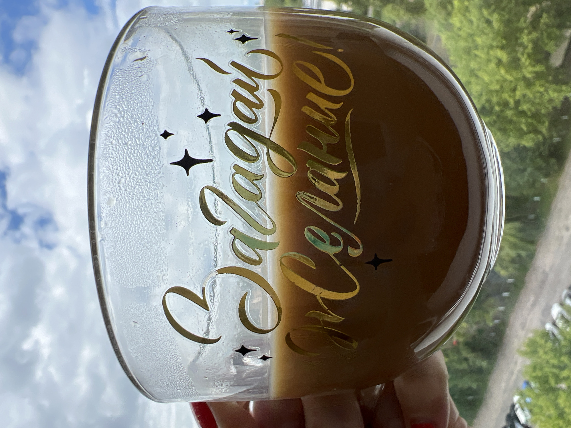 This media file shows a close-up, angled shot of a clear glass, likely a mug, filled with a dark brown beverage, possibly coffee or tea. The glass is decorated with gold, cursive lettering and small star shapes. The text reads "Загадай желанье," which translates from Russian to "Make a wish." The glass is partially fogged on the outside, suggesting it held a cold beverage or was exposed to a humid environment.
The background is blurred, showing a daytime scene with a bright, cloudy sky and a hint of green foliage and trees, indicating an outdoor setting. The reflection on the glass also suggests it's positioned near a window. A hand with red nail polish is visible at the bottom, holding the glass. The image is likely taken in Minsk, Belarus, given the context provided. The overall impression is one of a cozy, hopeful moment, perhaps enjoying a drink while contemplating a wish. The lighting suggests it's daytime, with natural light illuminating the scene.