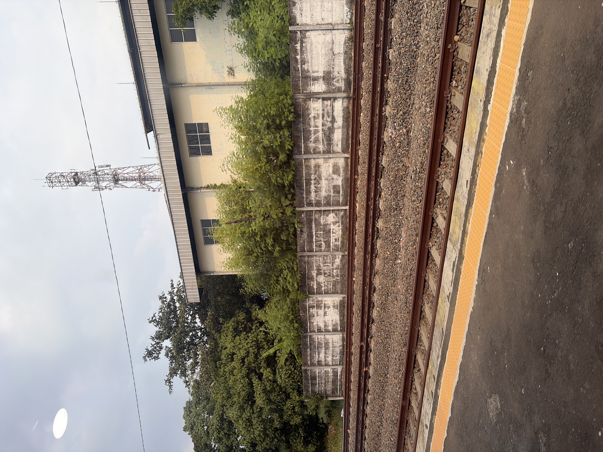 An outdoor, slightly hazy, overcast daytime scene shows a section of a train track. The foreground features a dark grey platform with a textured yellow safety strip along its edge, bordering the train tracks. The tracks themselves are comprised of gravel ballast, wooden ties, and metal rails, with multiple sets of parallel tracks visible. Beyond the tracks, a low, weathered concrete wall runs horizontally across the frame, exhibiting signs of age and discoloration. Behind the wall, dense green foliage and trees are visible. Further back, a light-colored, long, rectangular building with multiple windows stands. Atop the building, a tall, latticed telecommunications tower with antennas is prominent. A thin power line stretches across the upper portion of the frame. A large, bright, circular object, possibly a light fixture or lens flare, is in the upper left corner of the sky.