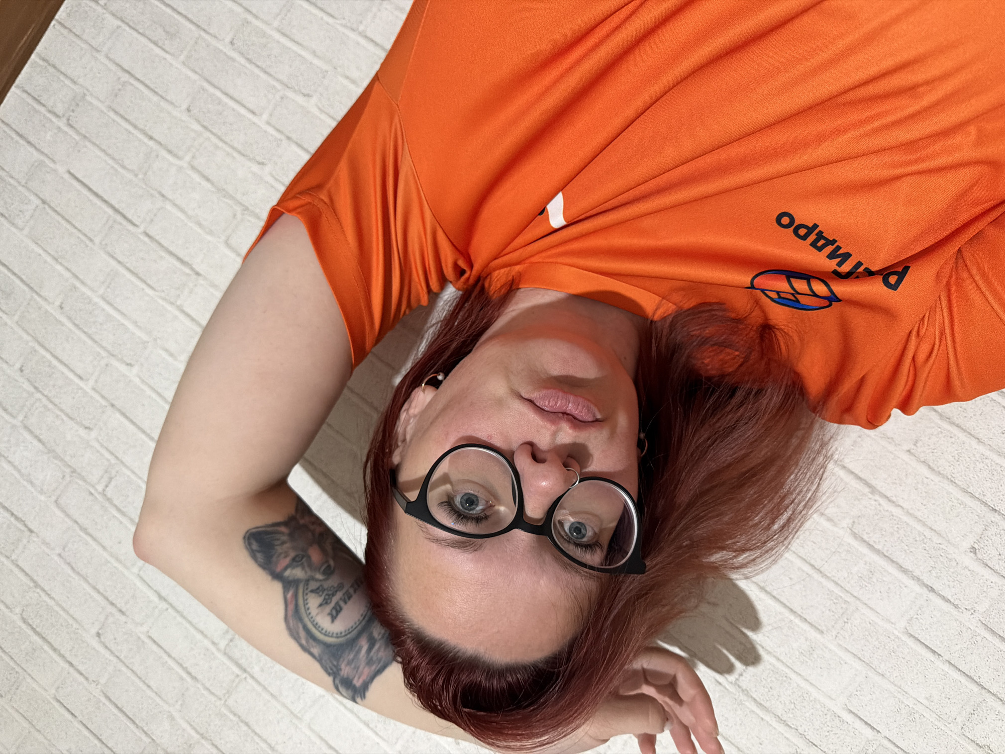 A Caucasian woman with red hair and glasses is lying on a white brick-patterned surface. She's wearing an orange T-shirt with the word "Рапидо" (Rapido) and a logo that appears to be a leaf with colored lines in blue and orange. She has a fox and clock tattoo on her left arm. Her head is tilted to the left, and she's looking directly at the viewer. She has a nose ring and earrings. The location may be in Chaikovsky, Russia, due to the Russian text on the shirt.