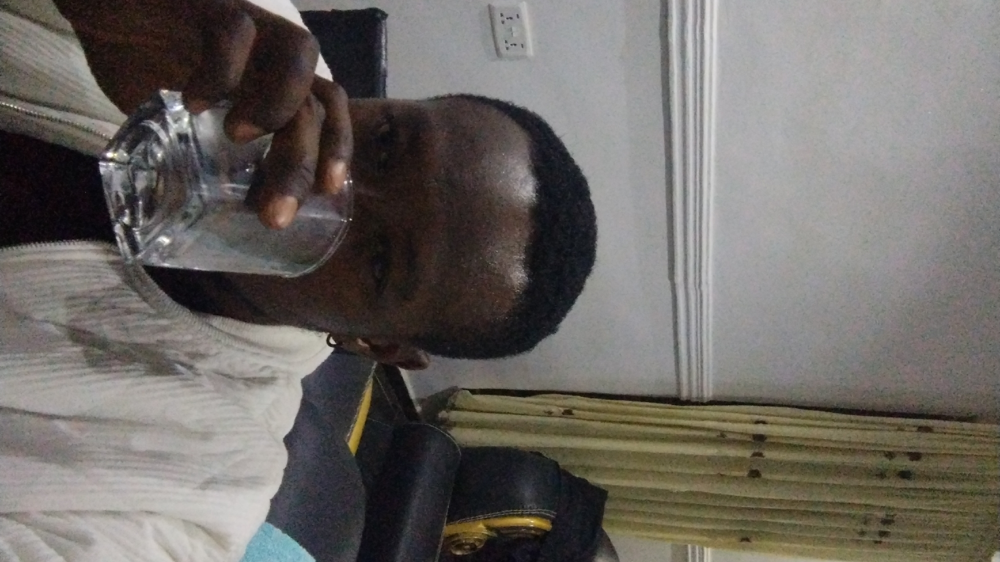 A person is drinking from a clear glass indoors. The person has dark skin and short, dark hair. They are wearing a white jacket with a zipper. The background includes a gray wall with molding, an electrical outlet, and wooden blinds.