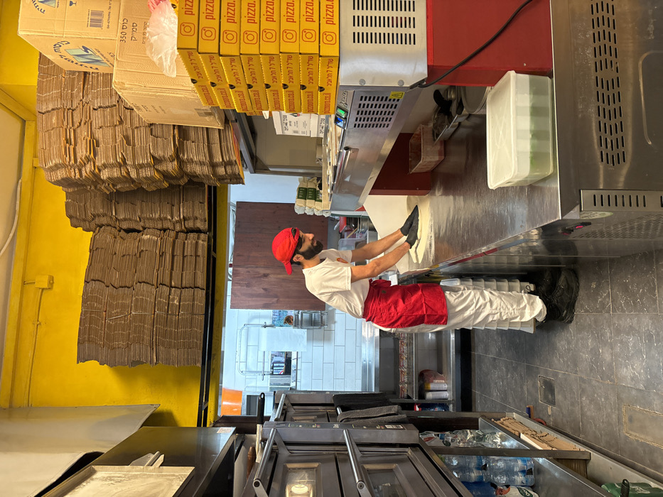 The image captures an interior scene within a commercial kitchen, specifically a pizzeria, located at Sderot 7 in Tel Aviv, Israel, as indicated by branding on the pizza boxes.
In the central foreground, a male individual, appearing to be in his late 20s to early 30s, is engaged in preparing pizza dough. He wears a red baseball cap, a white short-sleeved t-shirt with a logo, a red apron, white chef's pants, and black work boots. His hands are covered with black gloves as he actively flattens a circular piece of dough on a floured, stainless steel countertop. His facial expression indicates focus with a slight smile.
The workspace is a long, stainless steel counter. To the immediate right of the individual, a stack of approximately eight yellow pizza boxes is present. These boxes feature the "pizza.co.il" domain, an Instagram logo, and Hebrew text translating to "Sderot 7 Tel Aviv." Above this stack, a larger, brown cardboard box is positioned. Further to the right on the counter, a clear plastic container with a white lid holds a green substance, and a brown plastic bin is also visible. The counter incorporates built-in equipment with ventilation grilles and a digital display.
To the left of the individual, a multi-tiered commercial oven or prover unit with glass doors and controls is vertically stacked. Shelves beneath this unit hold several plastic water bottles.
The upper portion of the background features high stacks of flattened brown cardboard sheets, presumably for pizza boxes, stored on an elevated yellow shelf that extends across the width of the room. The back wall behind the individual exhibits white subway tiles, a small window-like opening, and an orange bucket hanging. A dark wooden panel is visible further back, serving as a partition. The floor is tiled with dark grey square tiles. The lighting is bright and even, consistent with a commercial kitchen environment.