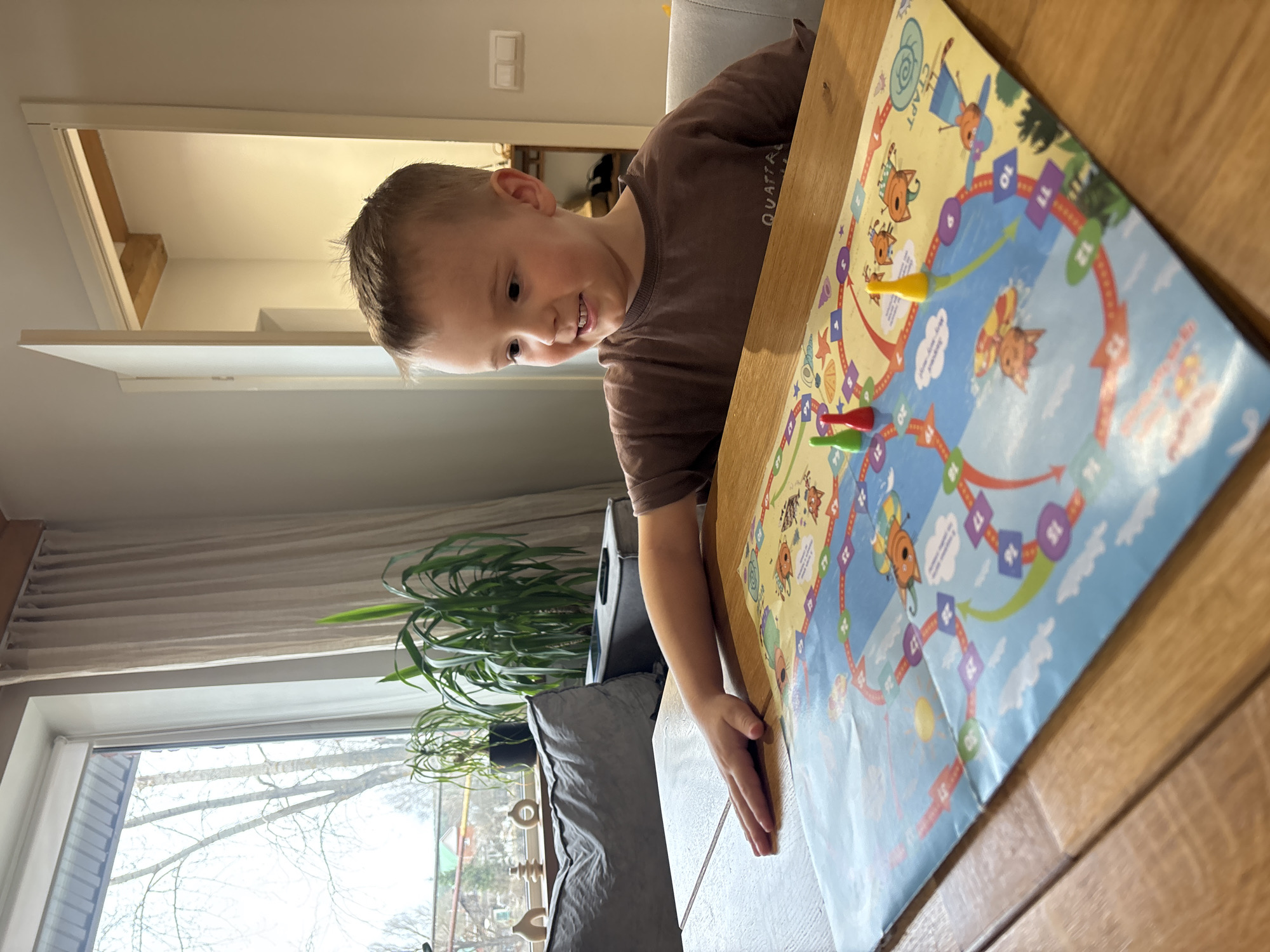 A child sits at a wooden table, engaging in a board game. The game board features a circular path with numbered spaces and cartoon animal illustrations. Three game pieces, colored red, green, and yellow, are positioned on the board. The child, wearing a brown t-shirt, gazes at the game with a slight smile. A window is visible in the background, showing a view of bare trees outside, indicative of winter or early spring. A plant sits near the window, and neutral-colored curtains hang above it. An open doorway is also visible, leading into a brightly lit room. No information to identify the city of Chaikovsky, Russia.