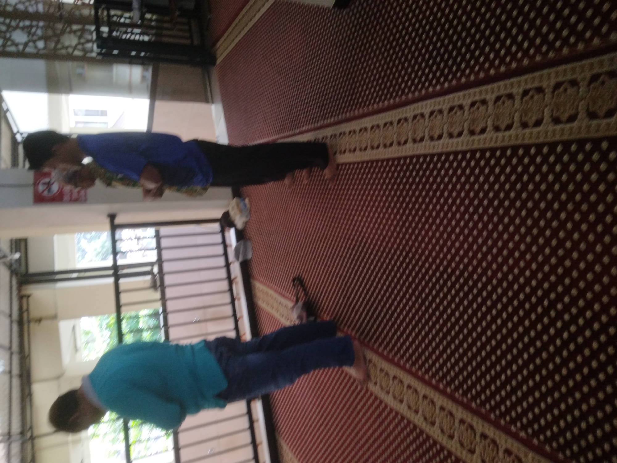 In Desa Pamijen Kecamatan Sokaraja, Indonesia, two men are standing on a patterned red carpet inside a building. The man on the left, facing away from the camera, is wearing a teal sweater and blue jeans. He is barefoot. The man on the right, also facing away from the camera, is wearing a blue t-shirt and black pants. He is also barefoot. Another person is partially visible behind and to the left of the man in the blue t-shirt. The background includes a railing, windows with greenery visible outside, and part of a wall. The lighting suggests an indoor environment, possibly a mosque or community center.