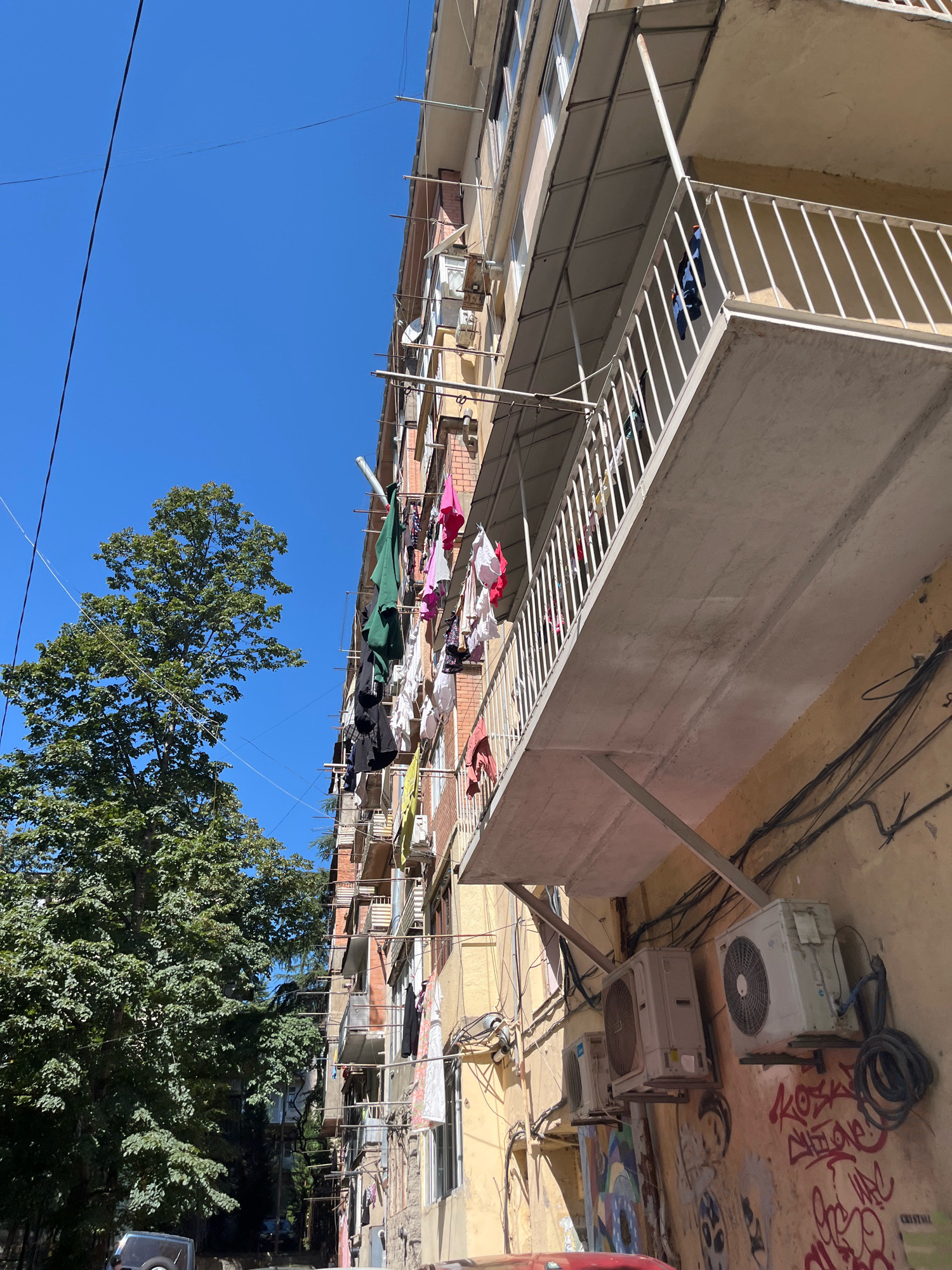 The image shows a low-angle view of a multi-story apartment building on a clear, sunny day in Tbilisi, Georgia. Laundry is hanging on clotheslines strung across balconies and between windows on the building's facade. The building appears to be an older Soviet-era structure with a yellowish-tan exterior. Several air conditioning units are mounted on the exterior wall, and there is graffiti visible at the base of the building. A large, leafy green tree is visible to the left of the building, and power lines are strung across the sky. A portion of a car is visible in the lower left corner of the image. The scene depicts a common urban residential setting in Tbilisi.
