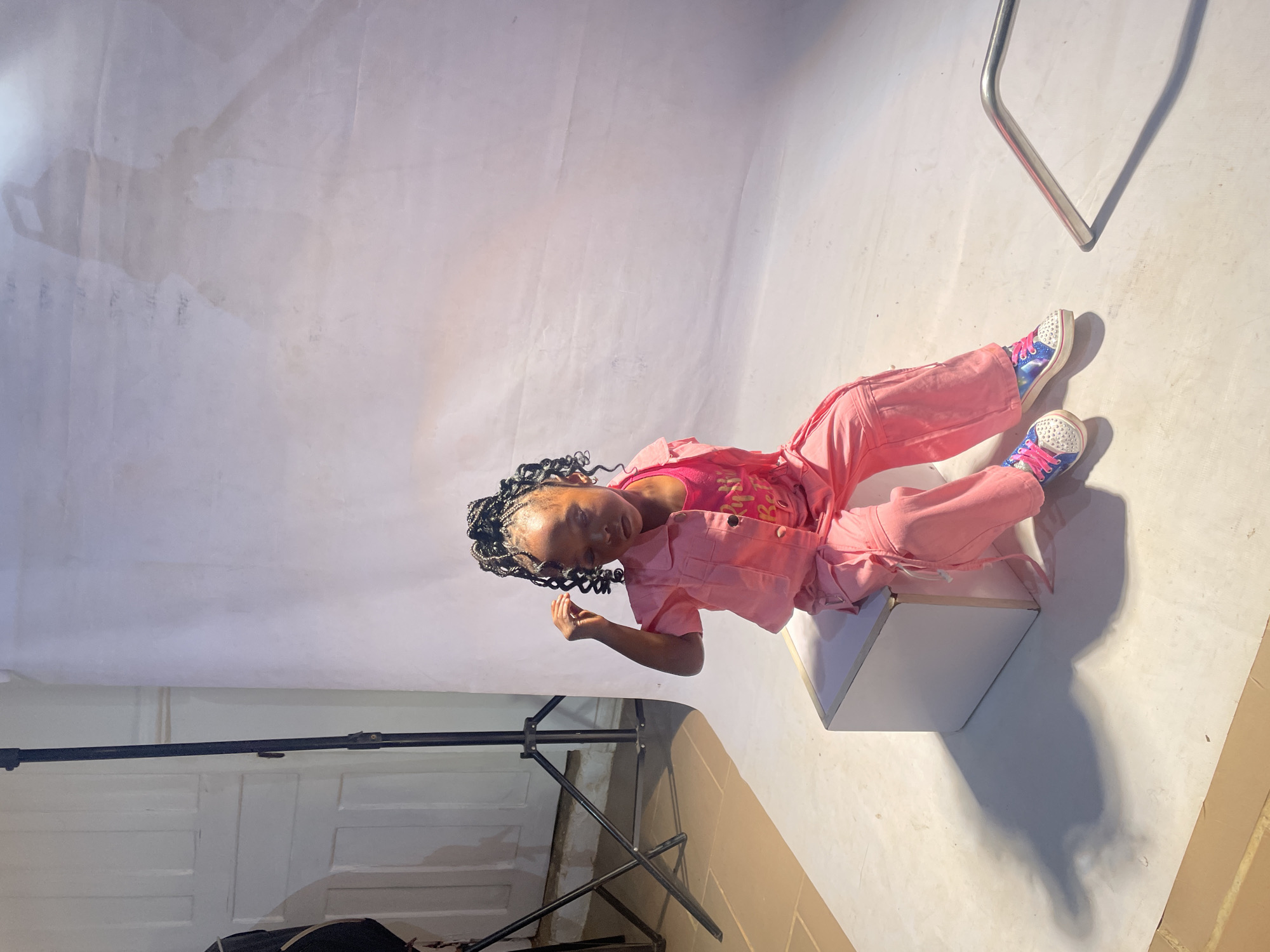 A young person with braided hair, wearing a pink outfit and shoes with a sparkly design, is posed sitting on a small white box in what appears to be a photography studio. The studio has a white backdrop, with lighting equipment and other studio paraphernalia visible in the background. The scene is likely in Barakin Akawo, Nigeria.