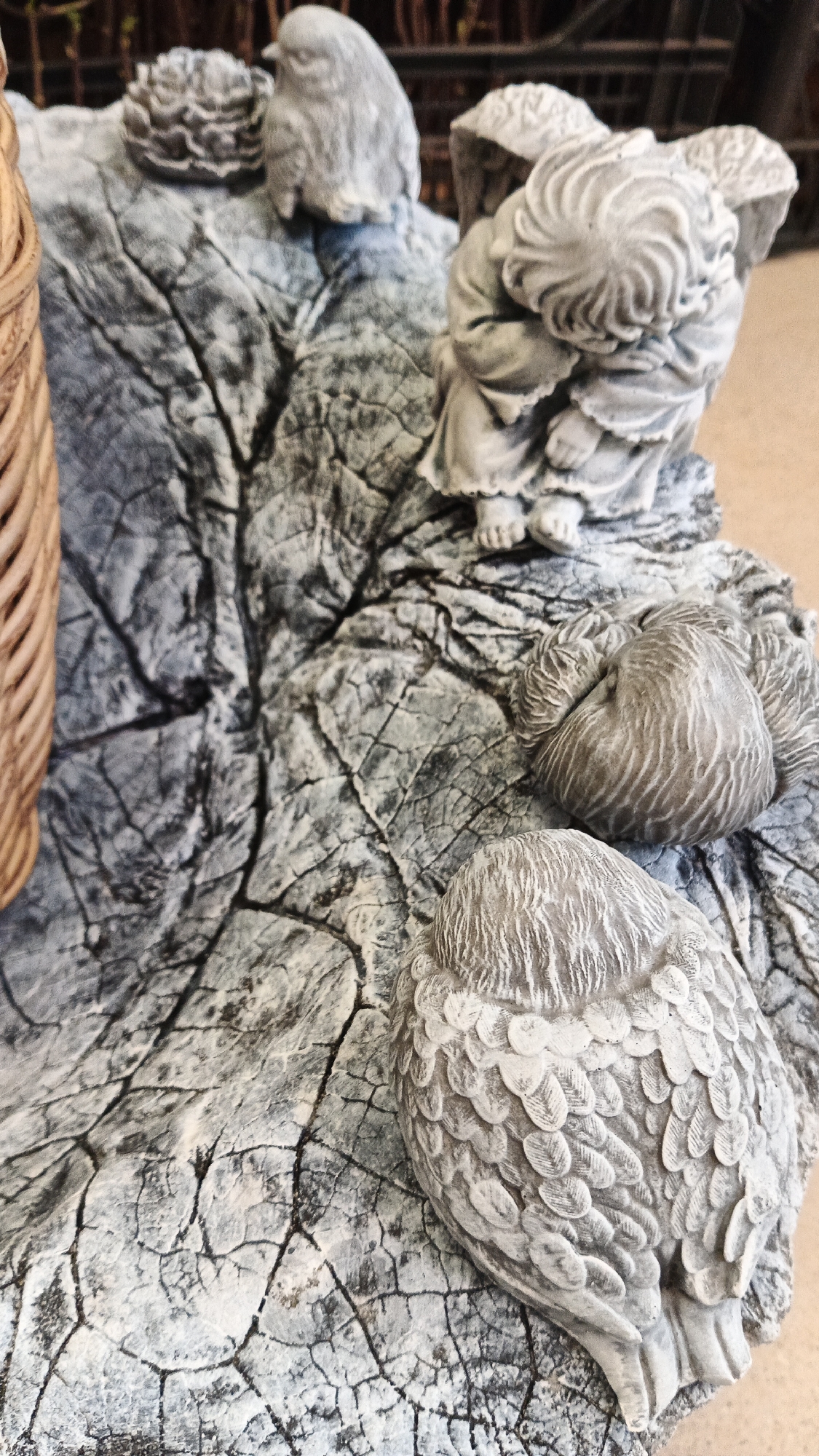 The image shows a close-up of a decorative garden statue, possibly made of concrete or resin, designed to resemble a weathered tree stump or rock formation. The statue features several small, sculpted figures. In the upper left, a small bird is perched on a textured surface. To its right, a winged figure, possibly an angel or cherub, is depicted in a seated or crouching position, with its head bowed. Further down and to the right, two more bird-like figures are shown, their heads tucked into their bodies. The entire scene is set against a textured, grey surface that mimics aged wood or stone, with visible cracks and grain patterns. A portion of a woven object, possibly a basket or decorative element, is visible on the far left edge of the image. The background is blurred, showing hints of greenery and possibly other garden items. There is no information in the image to suggest a specific location in Kryvyi Rih, Ukraine.