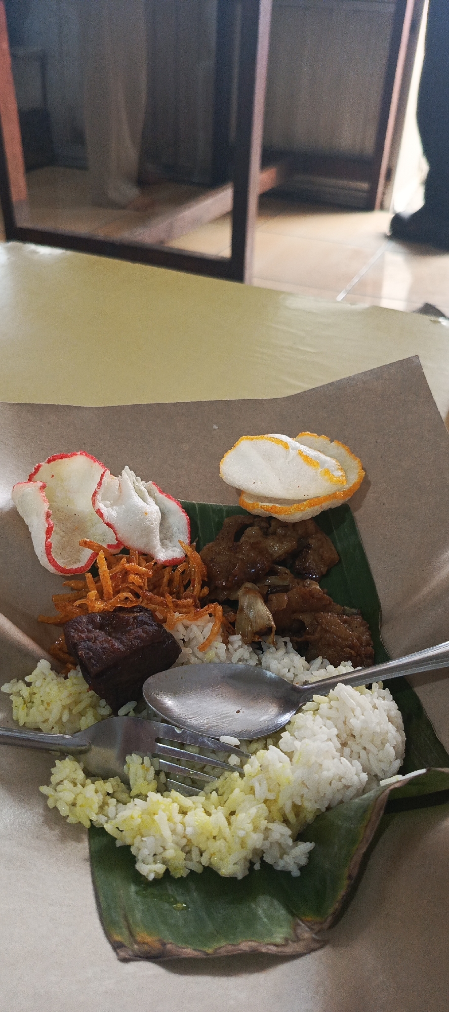 A meal served on a banana leaf with a portion of white rice, a portion of yellow rice, fried tempeh, shredded carrots, and crackers is displayed on a brown paper wrapper. The meal is on a table with a yellow tablecloth. The background is blurred, showing part of a wooden structure and people in dark clothing. This scene is likely in a restaurant or food stall in Sukaresmi, Indonesia.