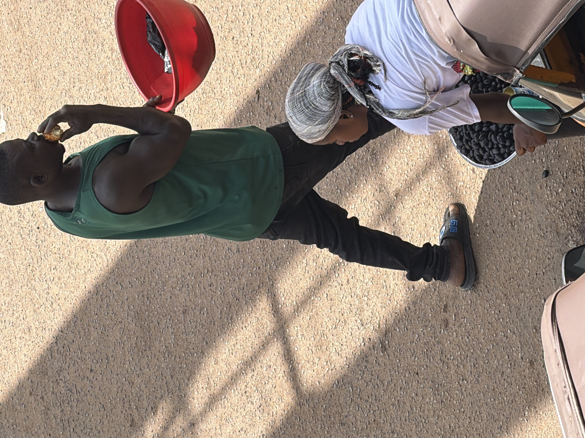 In Bukuru, Nigeria, a man wearing a green tank top and black pants walks while eating a piece of food, carrying a red bowl containing black plastic. Beside him, a woman in a white t-shirt and headscarf kneels, holding a bowl filled with dark, small, round items, possibly olives or dates, and leaning towards a three-wheeled vehicle. The ground is a light-colored, granular surface, with distinct shadows cast across it, suggesting a sunny day.