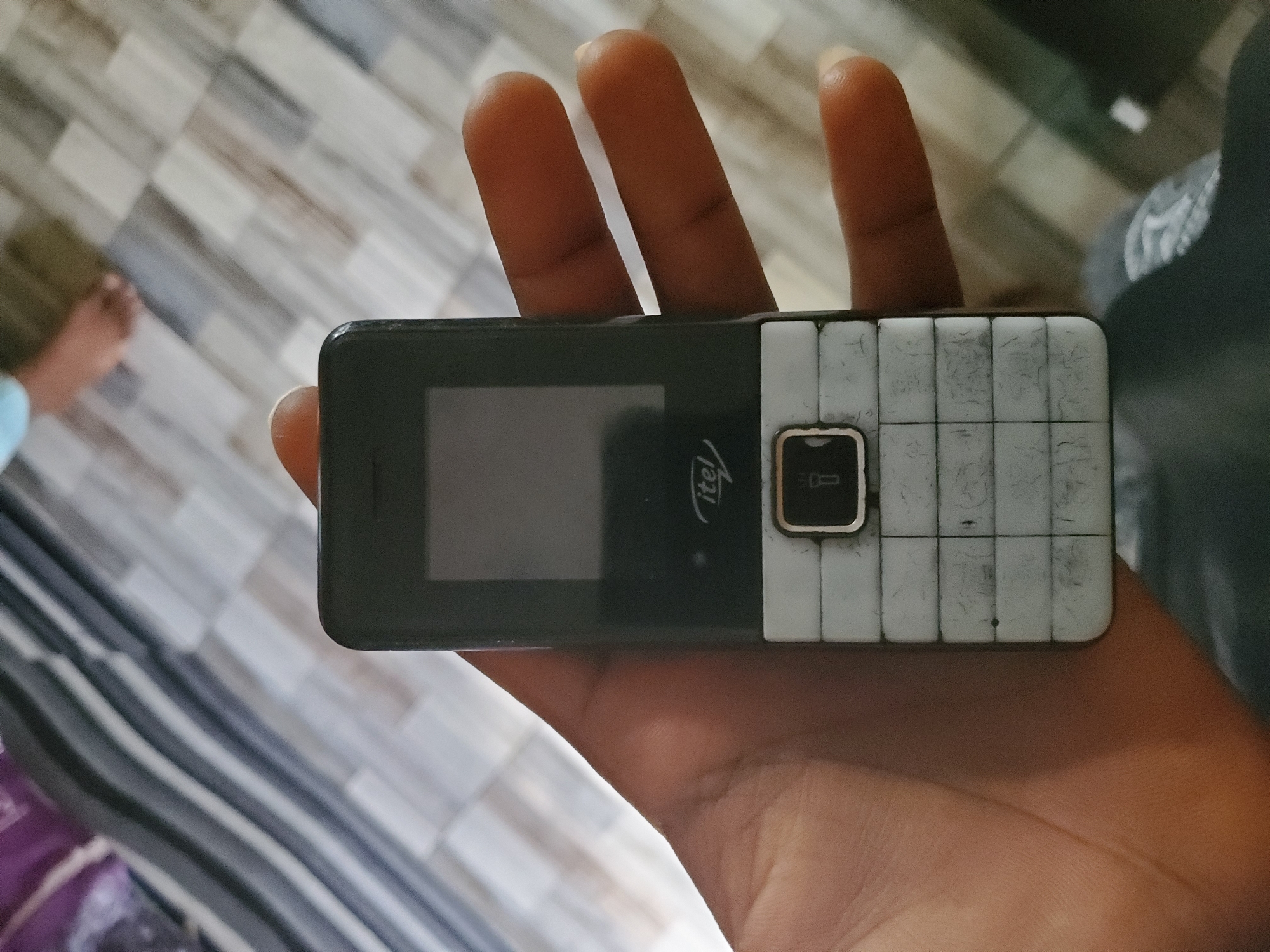 A hand holds a black and white Itel feature phone. The phone's screen is dark, and the keypad is composed of white buttons arranged in a grid. A raised button with a flashlight icon is visible on the keypad. The phone is positioned over a floor with a tiled pattern in shades of grey and beige. In the background, blurred figures and a striped fabric are visible. The scene is likely indoors, potentially a home or public space, in Jalingo, Nigeria.